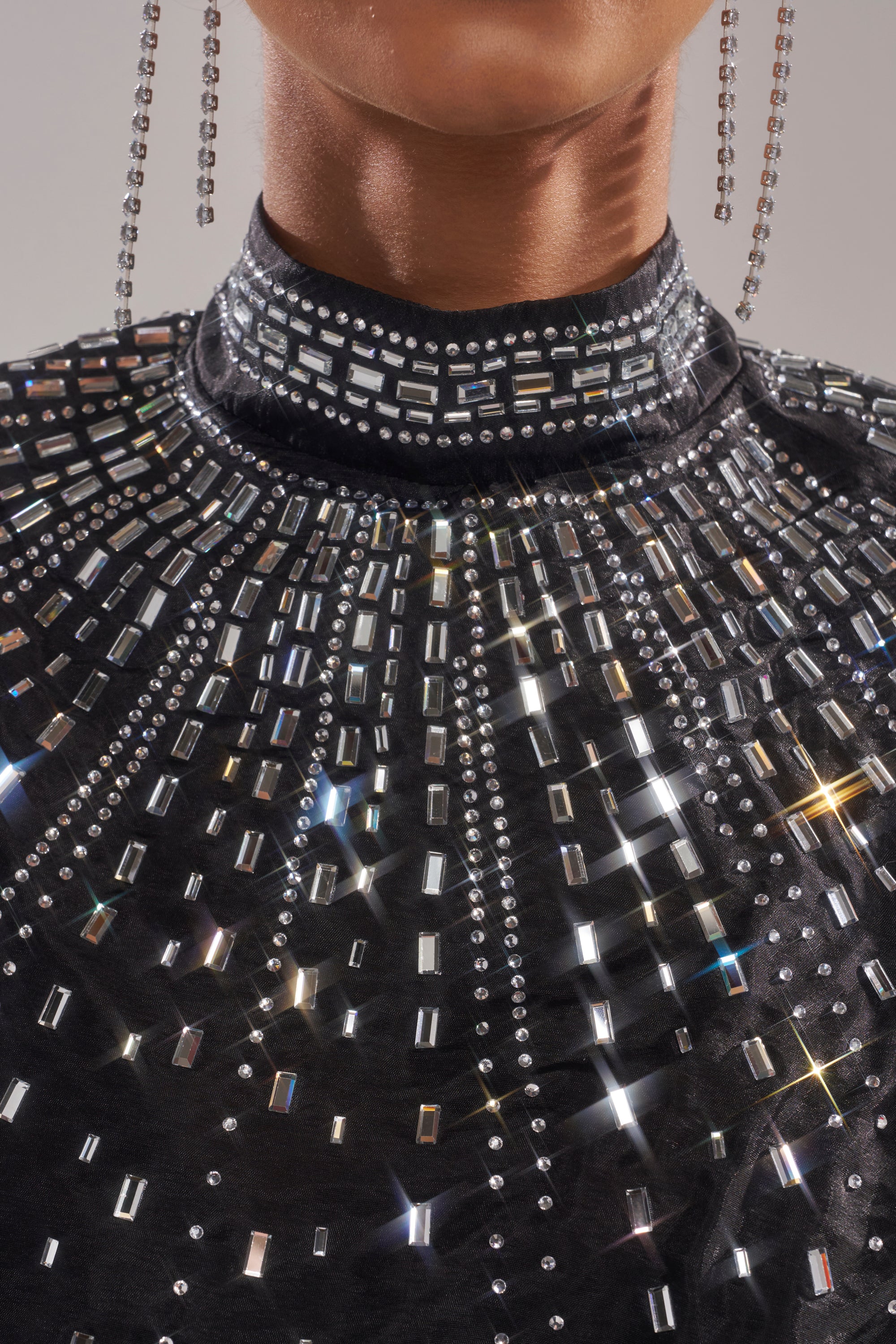 A close-up of a person wearing the GLAMOROUS RHINESTONE MOCK NECK BLOUSE—an elegant black high-neck top embellished with sparkling silver studs in a radiant, starburst pattern. Long, dangling earrings are also partially visible. 8
