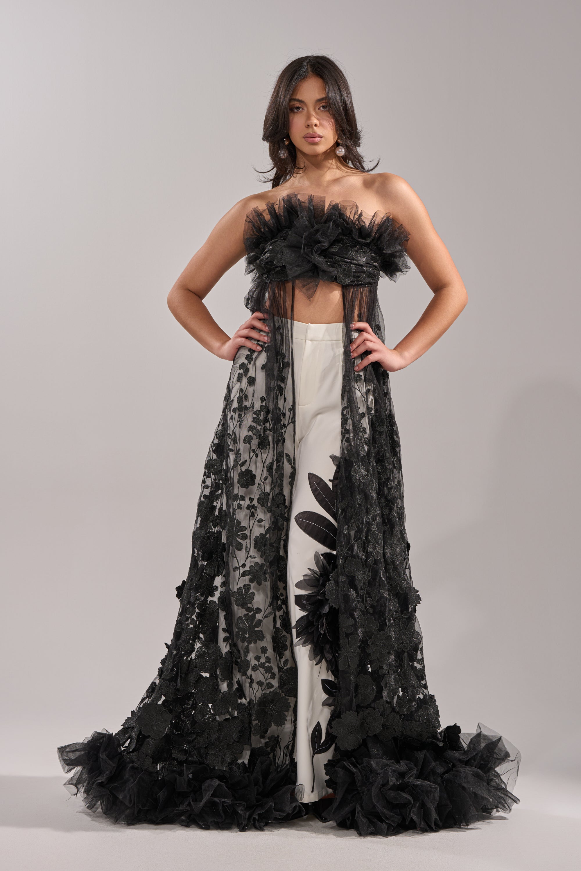 A woman exudes confidence in the DRAMA IS MY MIDDLE NAME BLOUSE—a black tulle strapless top with floral embroidery—paired with white pants featuring bold black floral prints. The sheer overlay and ruffled edges create a dramatic, elegant appearance. 0