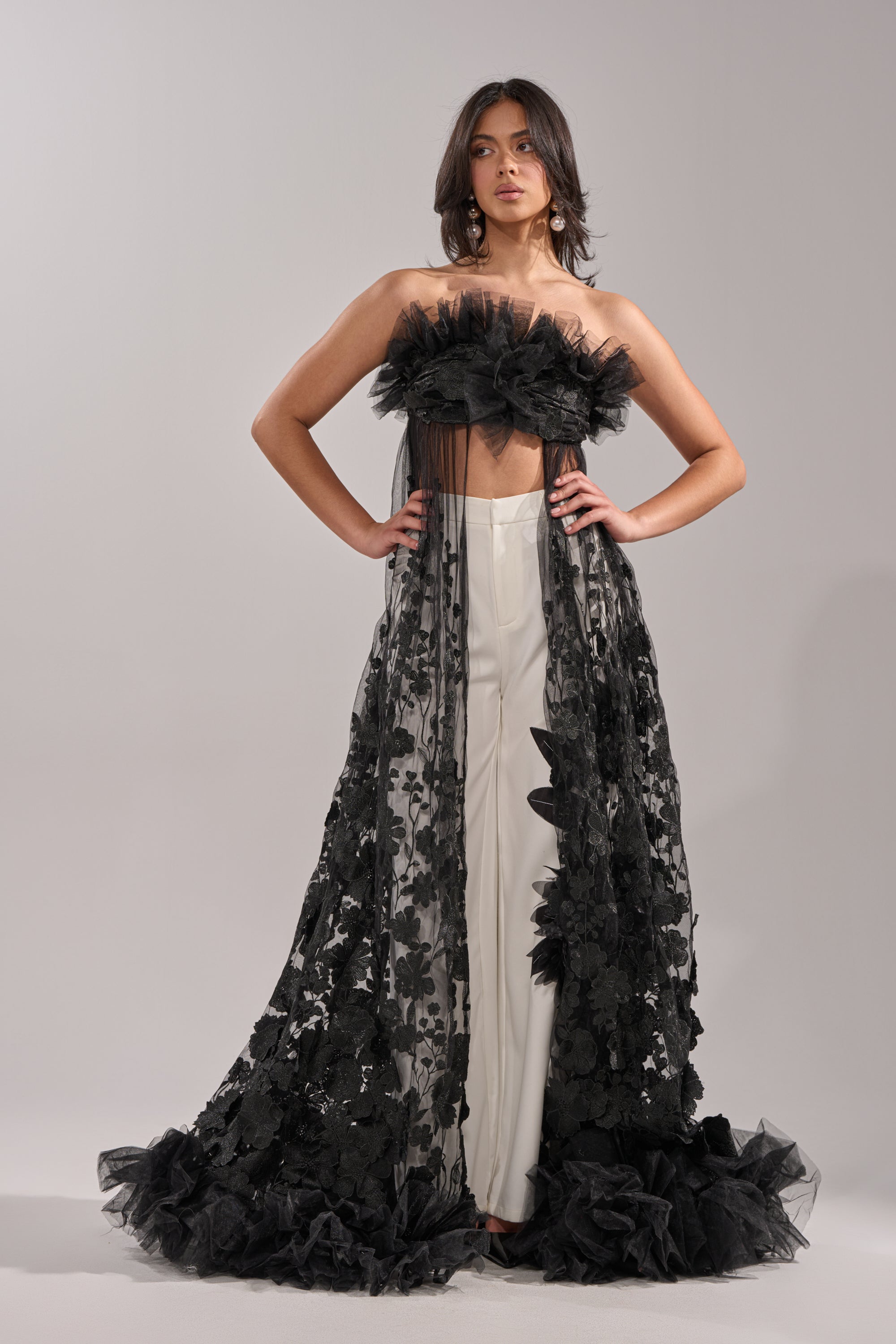 A woman exudes confidence in the DRAMA IS MY MIDDLE NAME BLOUSE—a floor-length, black tulle strapless top with floral embroidery, dramatic ruffles, and sheer fabric—styled over white pants as she poses hands on hips against a plain background. 4