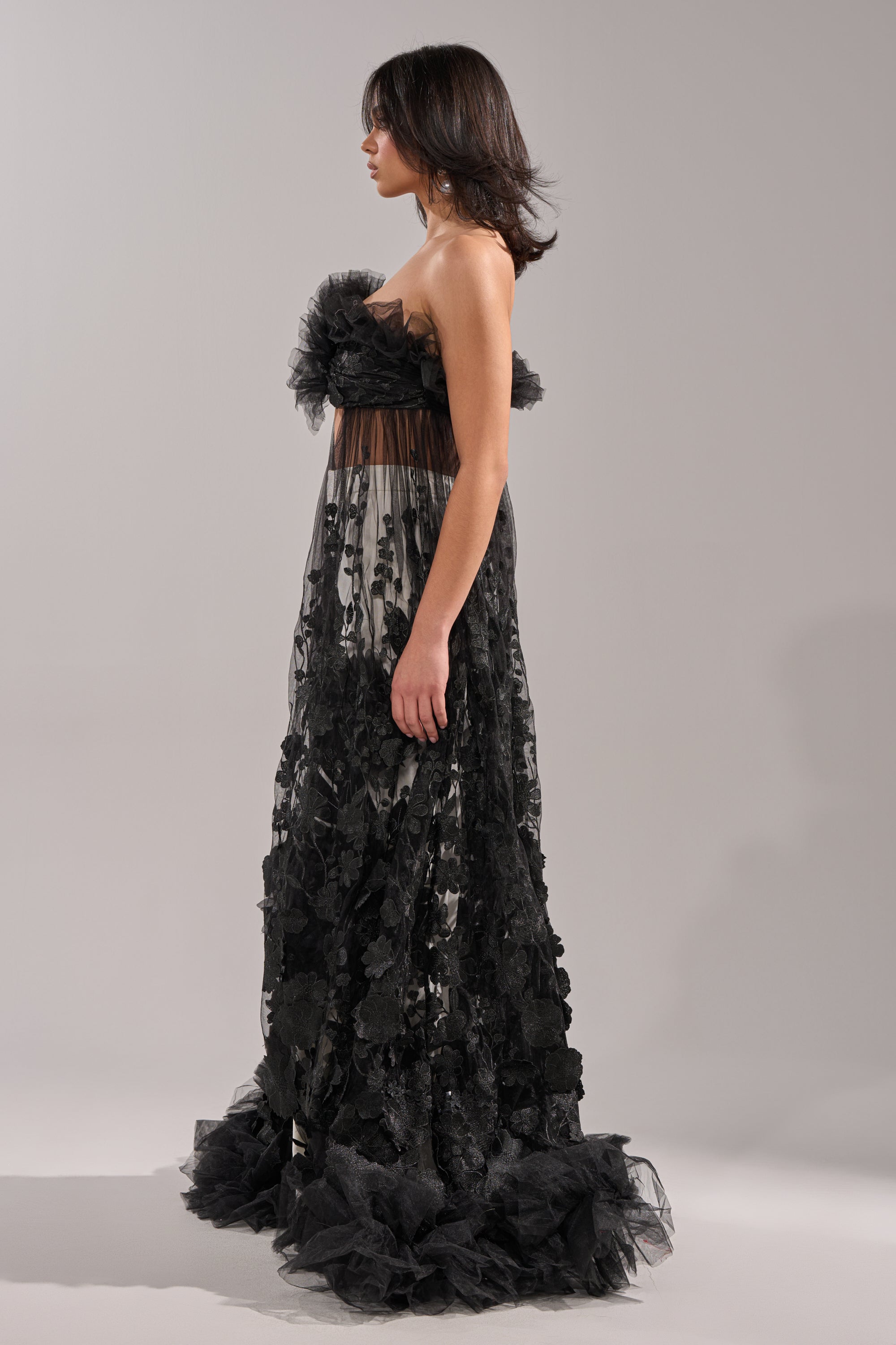 A woman stands in profile against a plain light gray background, wearing the DRAMA IS MY MIDDLE NAME BLOUSE—a black strapless top with sheer floral embroidery and ruffled tulle details, styled with floor-length evening wear. 5