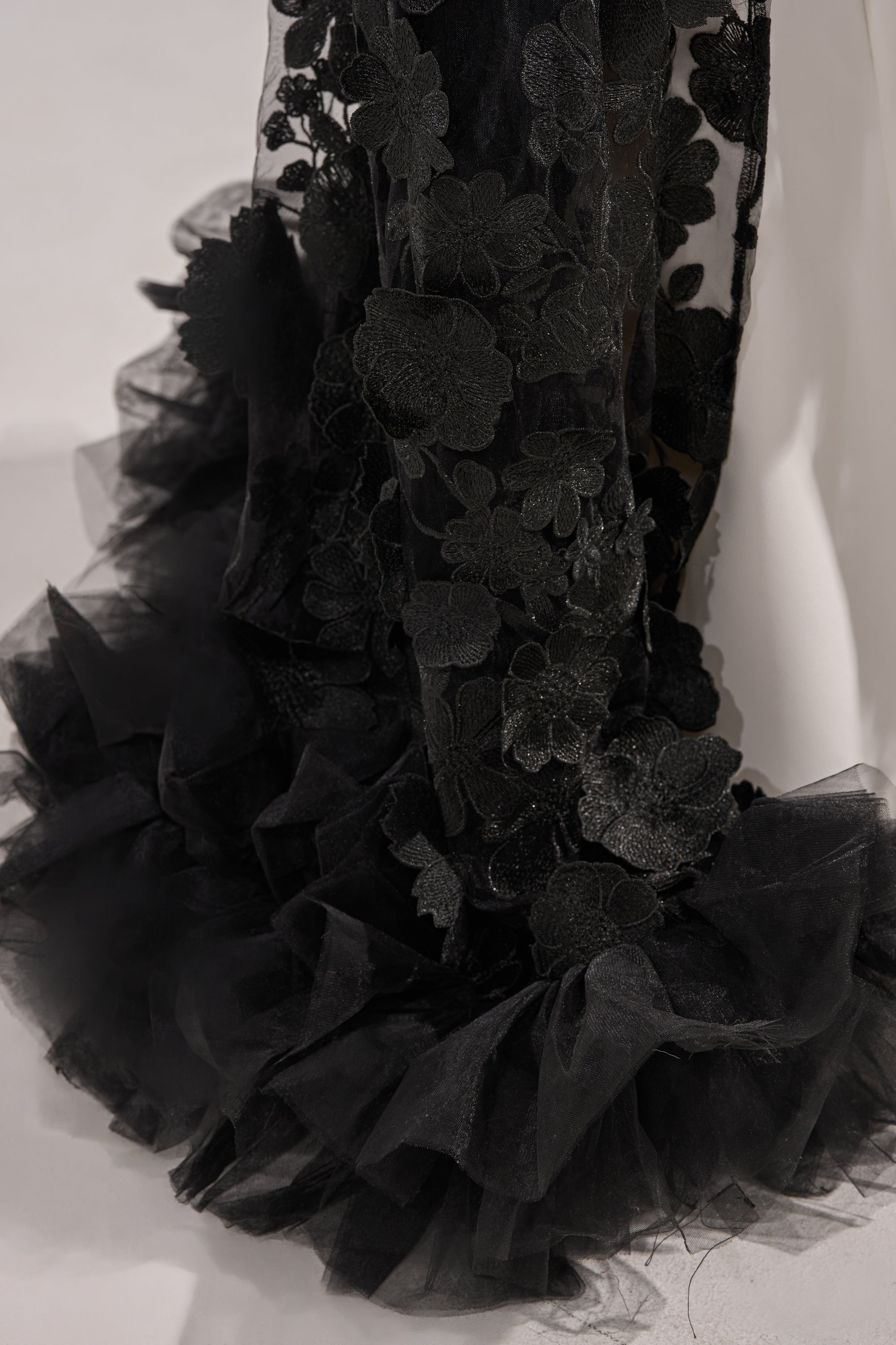Close-up of the DRAMA IS MY MIDDLE NAME BLOUSE in black sheer fabric with floral embroidery and a ruffled tulle hem, draping elegantly against a light background—ideal for striking evening wear or as a chic statement top. 8