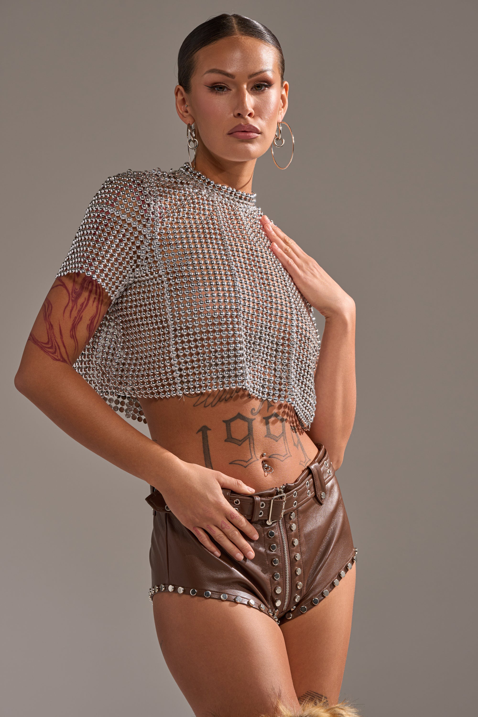 A woman poses confidently in the BEADS FOR DAYS TEE, a beaded crop top with short sleeves, paired with high-waisted brown leather shorts featuring studs and buttons. She completes the look with hoop earrings, visible tattoos, and a sleek pulled-back hairstyle. 0