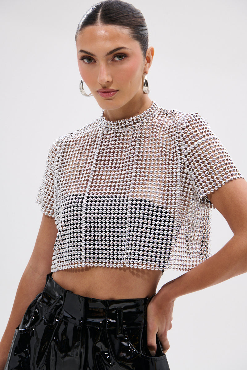 A woman with dark hair in a sleek bun wears the BEADS FOR DAYS TEE, a short-sleeve mesh top adorned with silver rhinestones and ball-chain details over a black crop top, paired with shiny black high-waisted pants.