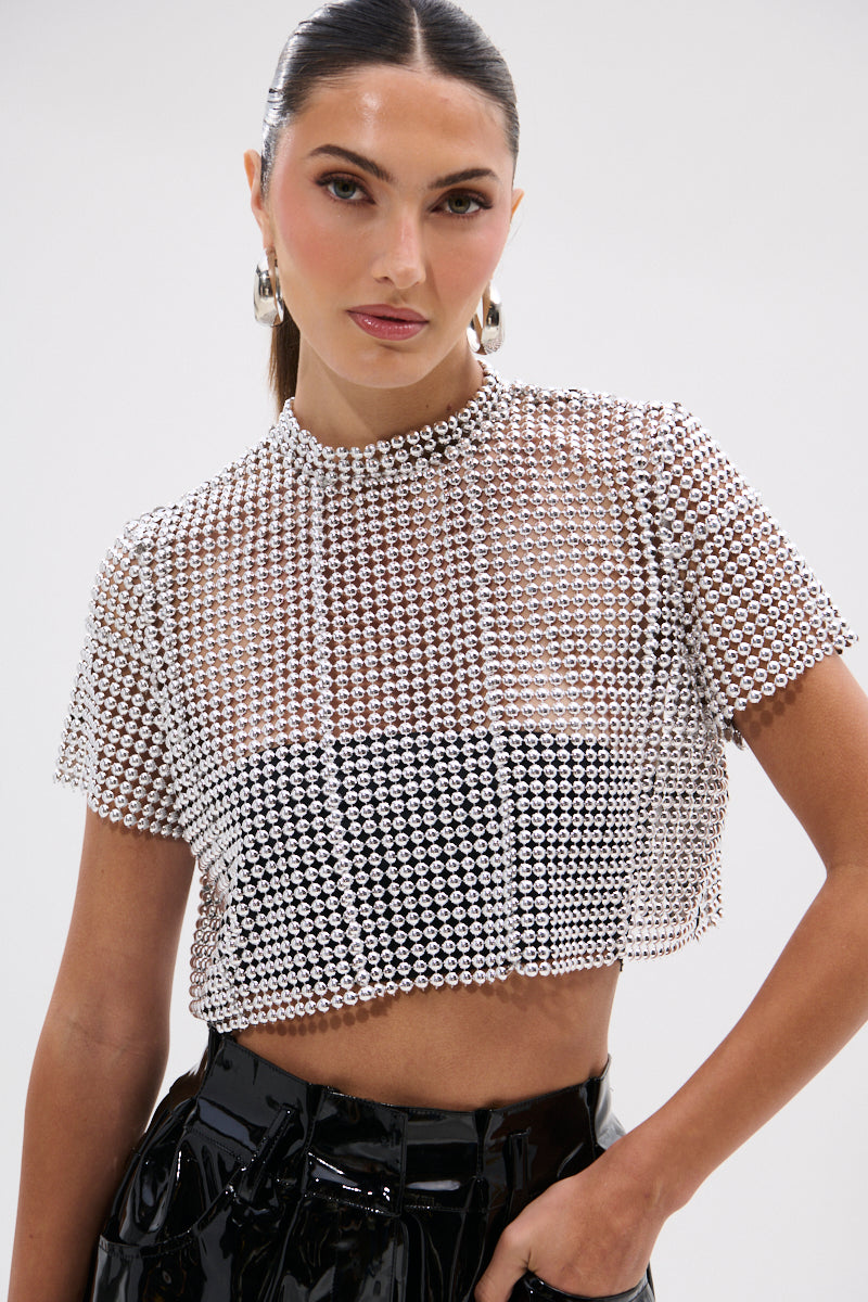 A woman with slicked-back hair wears the BEADS FOR DAYS TEE—a short-sleeve, high-neck beaded crop top with silver ball-chain embellishments—over a black bralette and glossy black pants, posing against a plain background.
