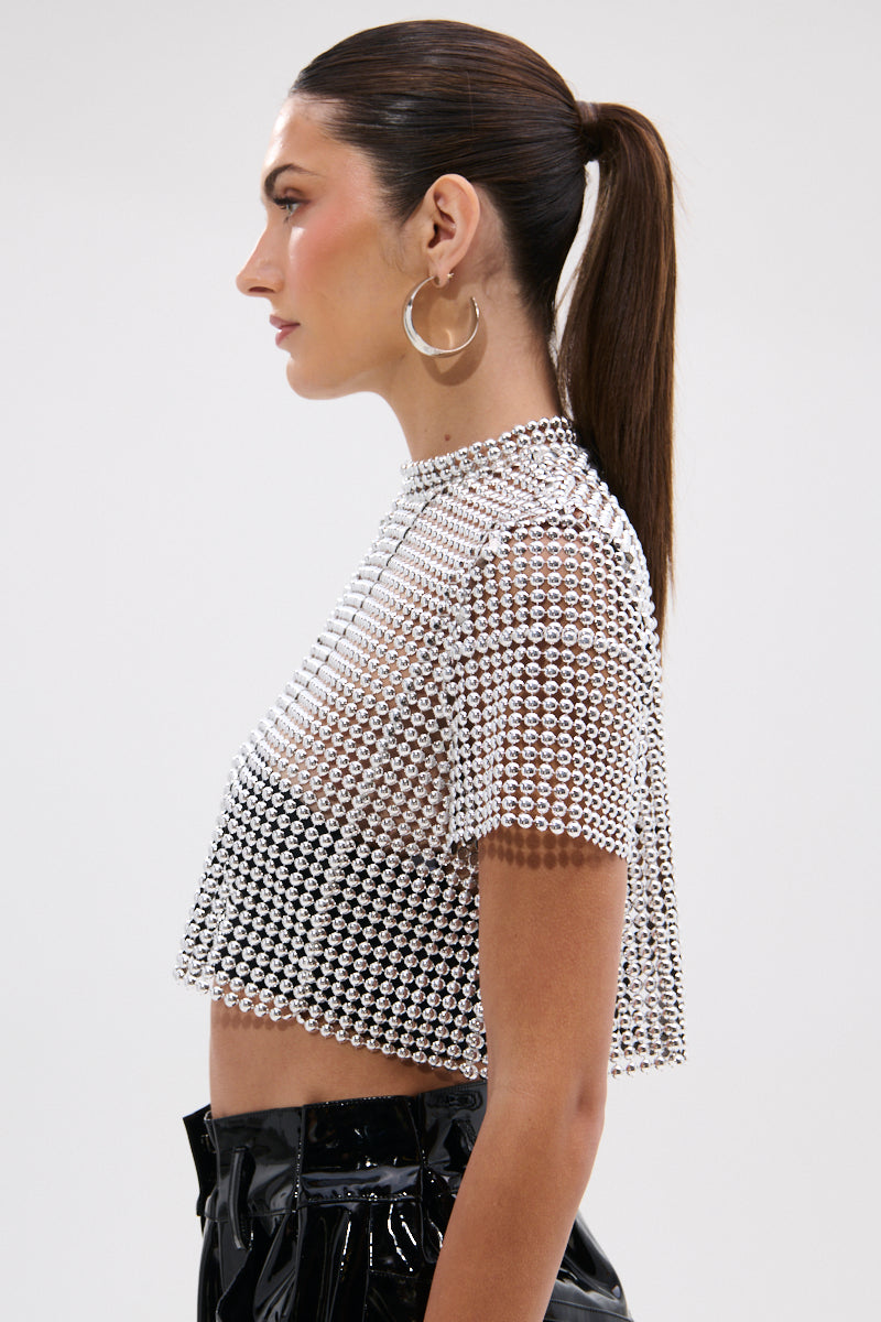 A woman with long brown hair in a ponytail models the BEADS FOR DAYS TEE—a sheer beaded crop top with silver ball-chain details—and shiny black high-waisted pants, styled with large hoop earrings, shown in profile against a plain white background.
