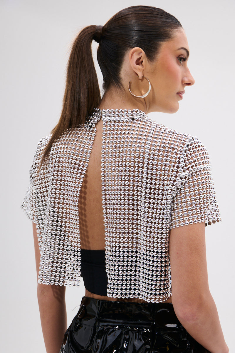 A woman with a sleek ponytail wears large hoop earrings and the BEADS FOR DAYS TEE—a short-sleeve, open-back beaded crop top with silver ball-chain details over a black bandeau—paired with shiny black bottoms.