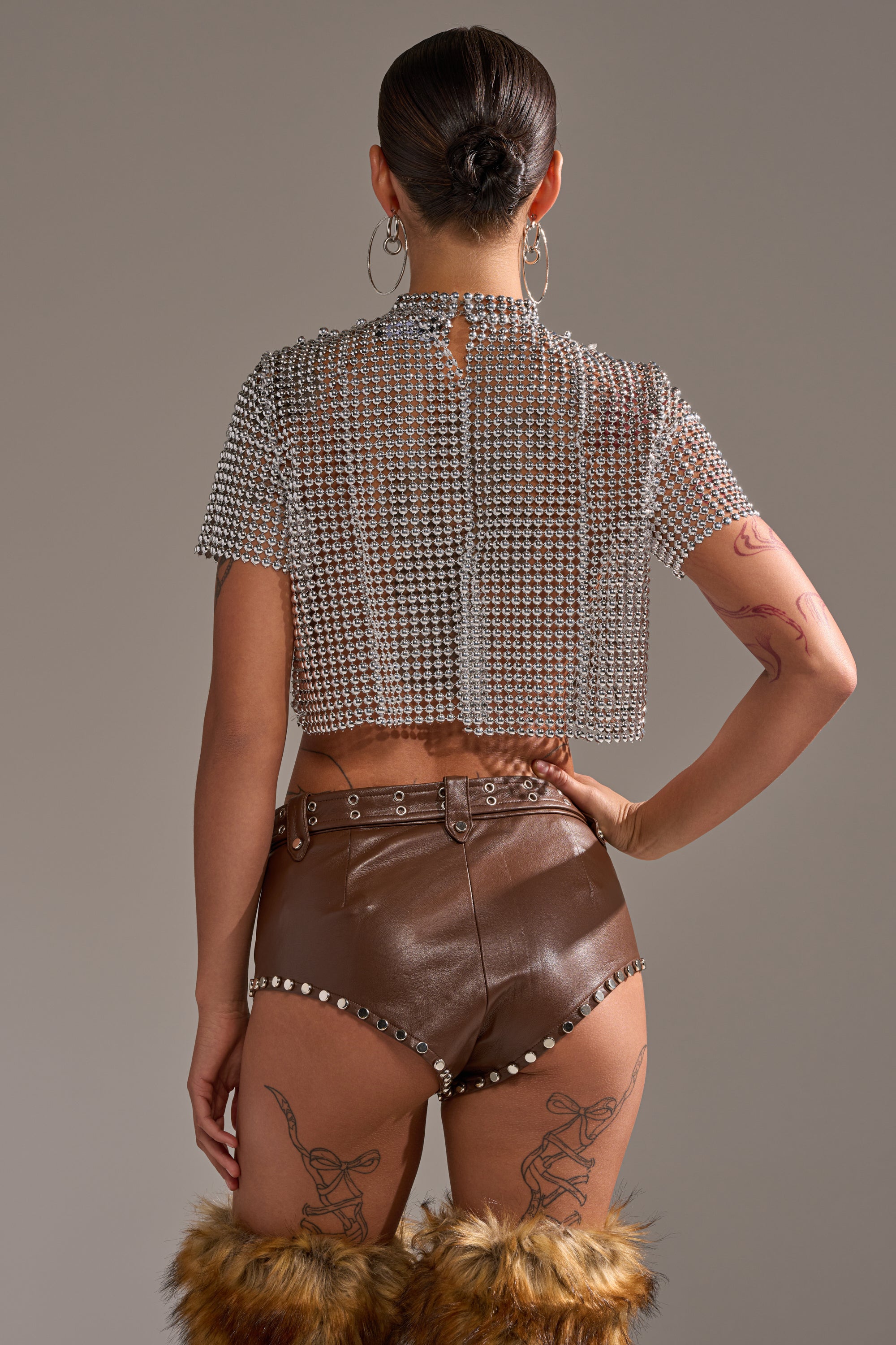 A person stands facing away, wearing the BEADS FOR DAYS TEE with an open-back design, paired with high-waisted brown leather shorts featuring silver studs and fur-trimmed boots. Their hair is in a bun, showcasing tattoos on their arms and legs. 4