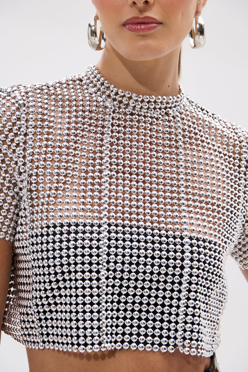 A woman wears the BEADS FOR DAYS TEE, a shiny silver beaded crop top with a high neckline layered over a black bandeau, paired with large silver hoop earrings and natural makeup.