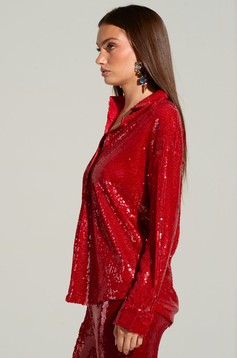 A woman with long, straight hair is wearing the vibrant red LOVER ZONE SEQUIN SHIRT from AKIRA Label, paired with matching pants. Her outfit gleams with intricate sequin detailing over a polyester base. Colorful dangling earrings add a touch of flair as she is photographed in profile against a neutral backdrop.