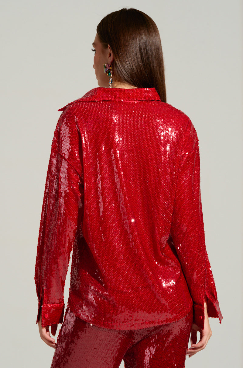 A person facing away, wearing the AKIRA LOVER ZONE Sequin Shirt along with matching pants. This shimmering red outfit, crafted from polyester and adorned with intricate sequin detailing, sparkles under the light. The individual has long brown hair and is positioned against a neutral background.