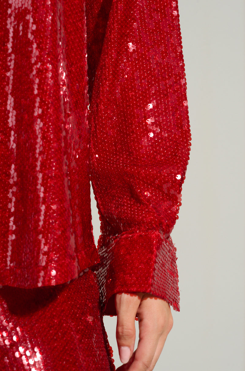 A close-up of a person wearing a dazzling red LOVER ZONE SEQUIN SHIRT. The focus is on the sleeve and upper part of the attire, showcasing tightly packed sequin detailing reflecting light. The hand is slightly visible, adding a touch of elegance to the polyester base ensemble.