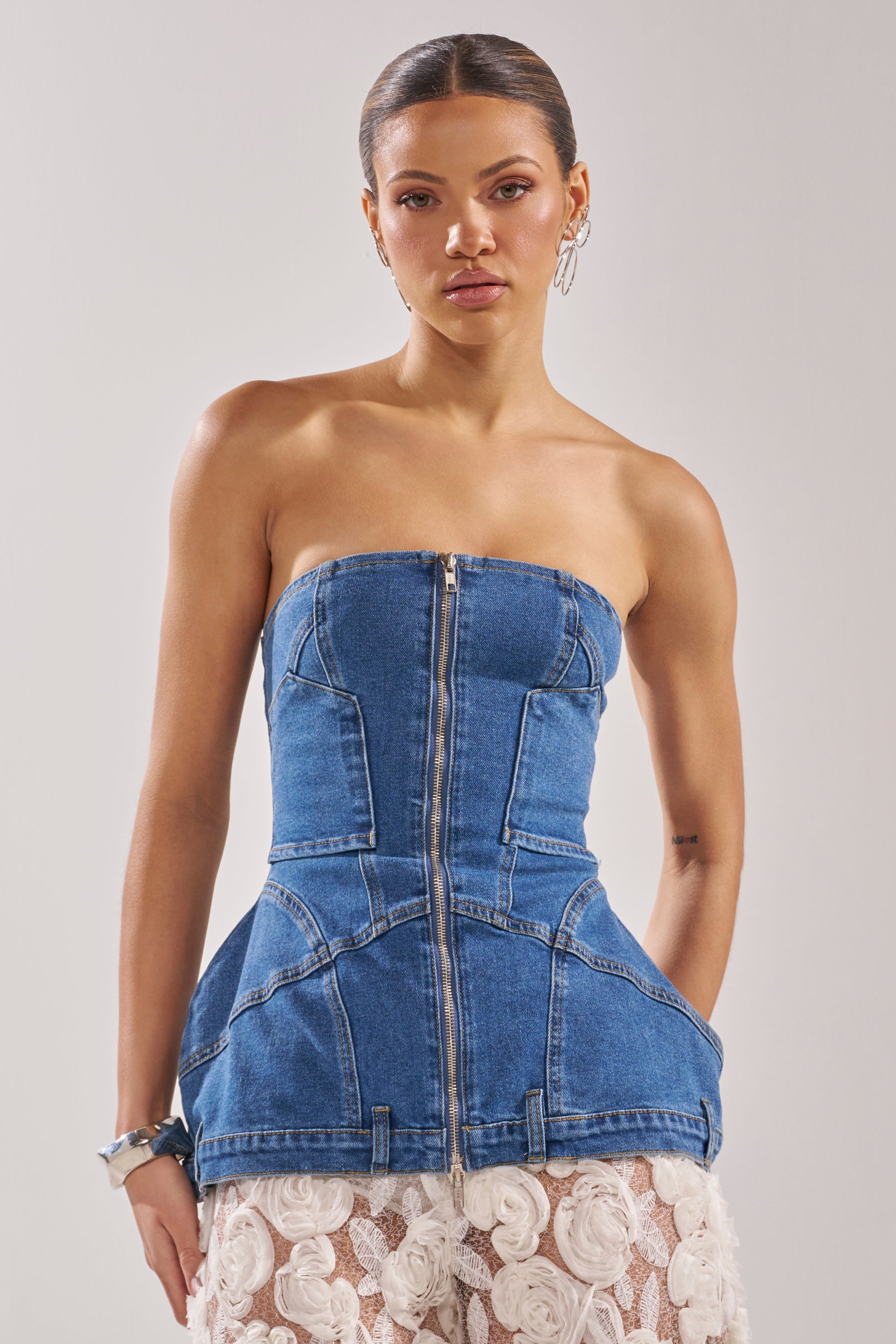 A woman with slicked-back hair wears the UPSIDE DOWN DENIM CORSET with a front zipper and peplum detail, paired with a white skirt featuring textured floral patterns, standing against a plain light background. 0