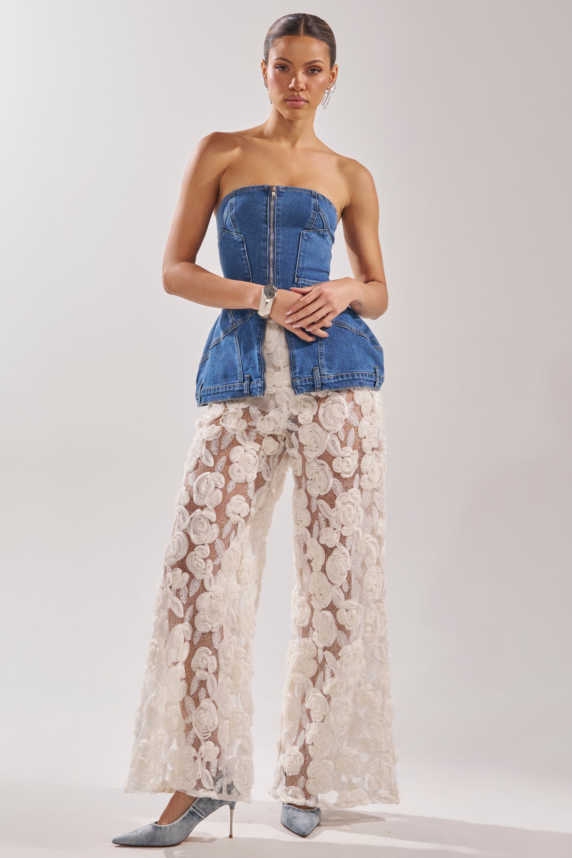 A woman models the UPSIDE DOWN DENIM CORSET, a strapless denim top with front zipper, styled with sheer white lace wide-leg pants and silver heels against a light background. 9