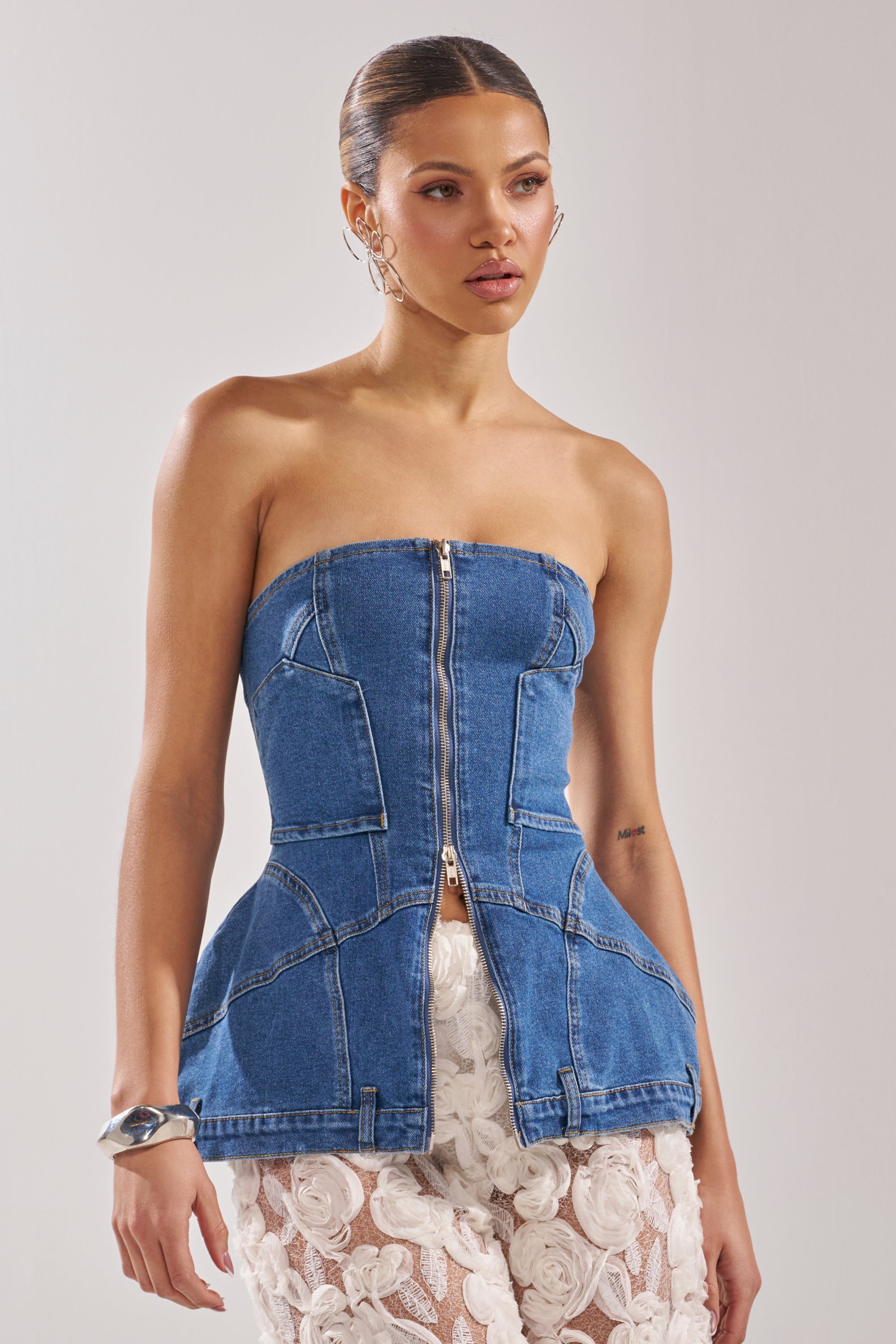 A woman wears the UPSIDE DOWN DENIM CORSET, a strapless denim top with a front zipper, paired with white floral lace pants, standing against a plain light background. 1