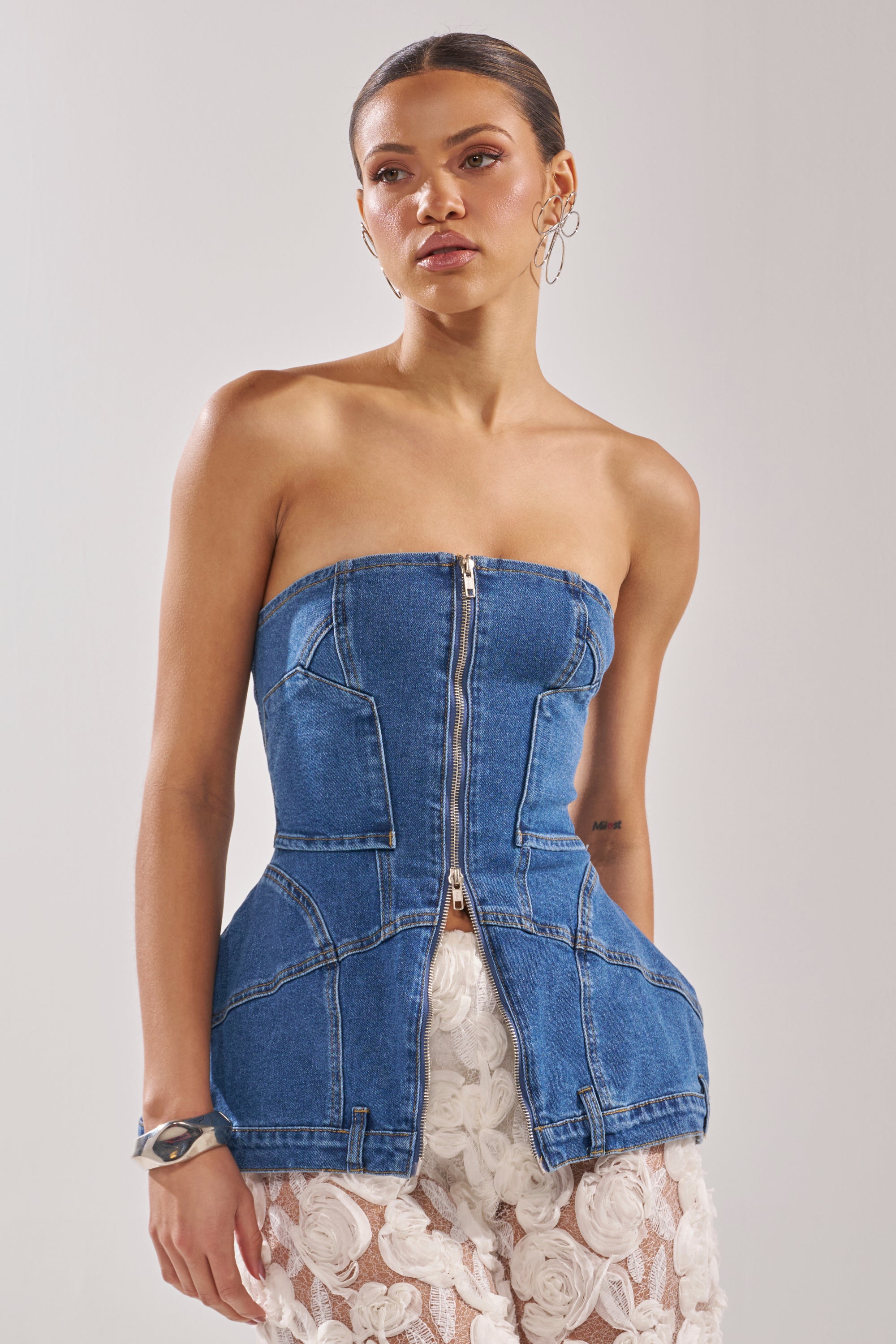 A woman models the UPSIDE DOWN DENIM CORSET, a strapless peplum denim top, paired with white lace-patterned pants, hoop earrings, and a chunky bracelet, posed against a plain light background. 2