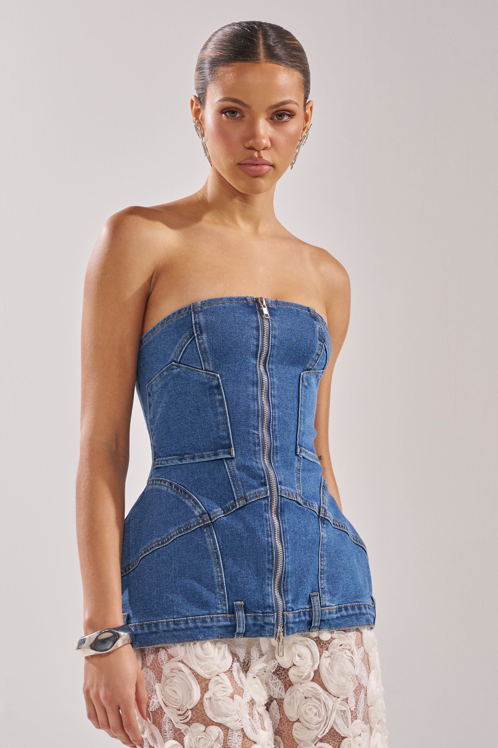 A woman models the UPSIDE DOWN DENIM CORSET—strapless with a front zipper and large pockets—paired with a white floral lace skirt, standing against a plain light background. 4