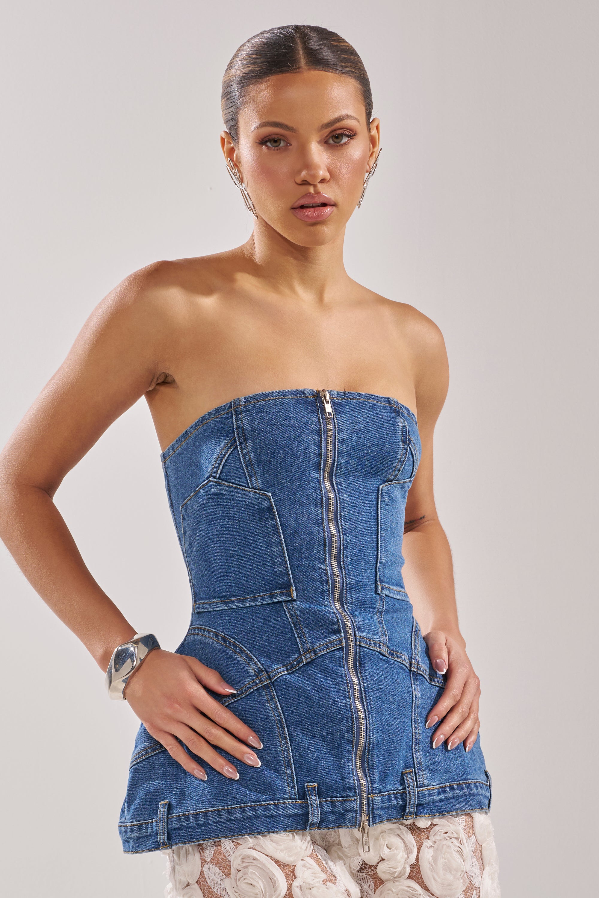 A woman models the UPSIDE DOWN DENIM CORSET—a strapless denim top with a zip front, visible seams, and pockets—styled with a chunky silver bracelet and statement earrings against a plain background. 5
