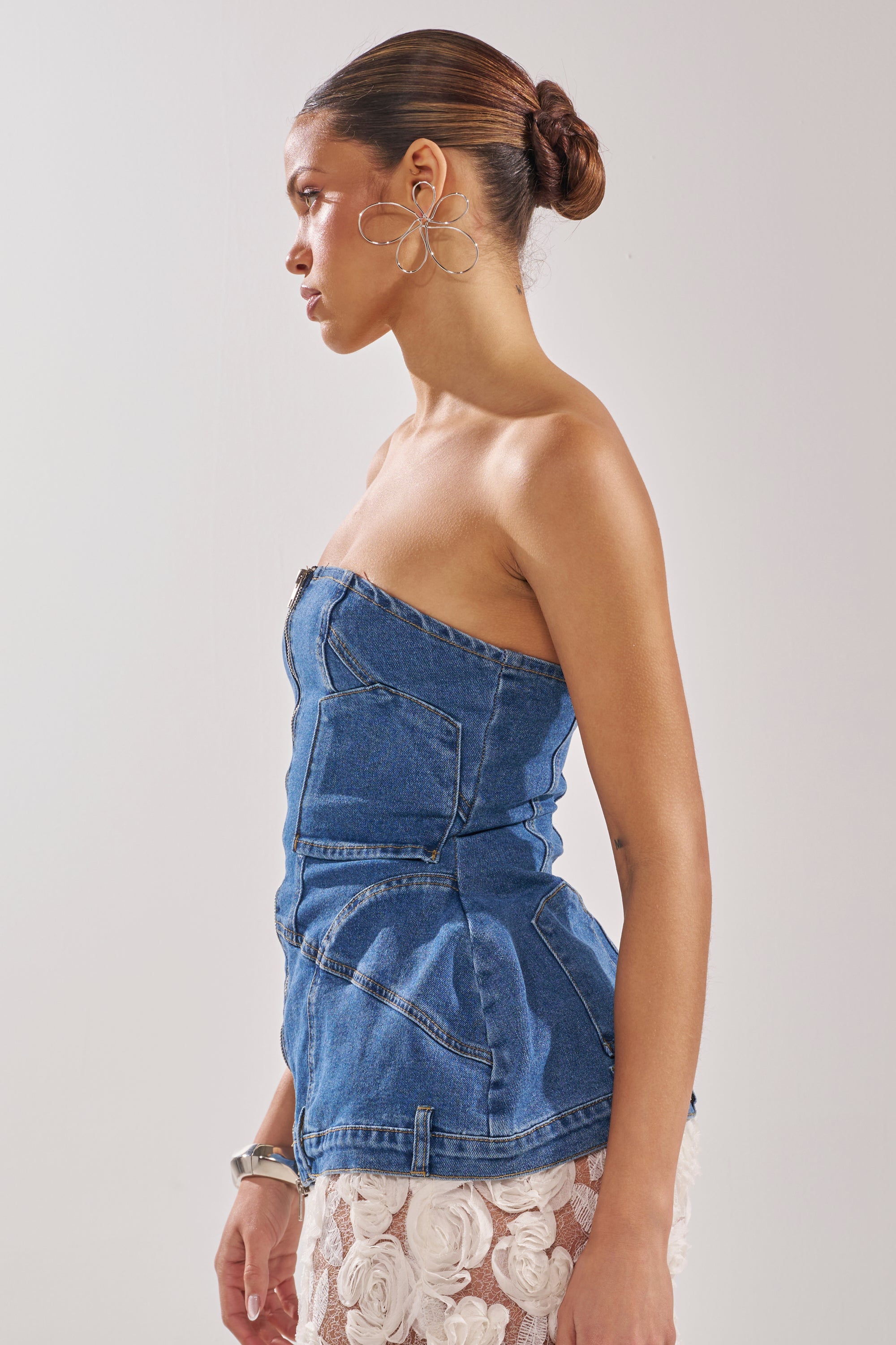 A woman with her hair in a bun models the UPSIDE DOWN DENIM CORSET, styled strapless with visible stitching, paired with a sheer white skirt, flower-shaped earrings, and a silver bracelet against a plain background. 6