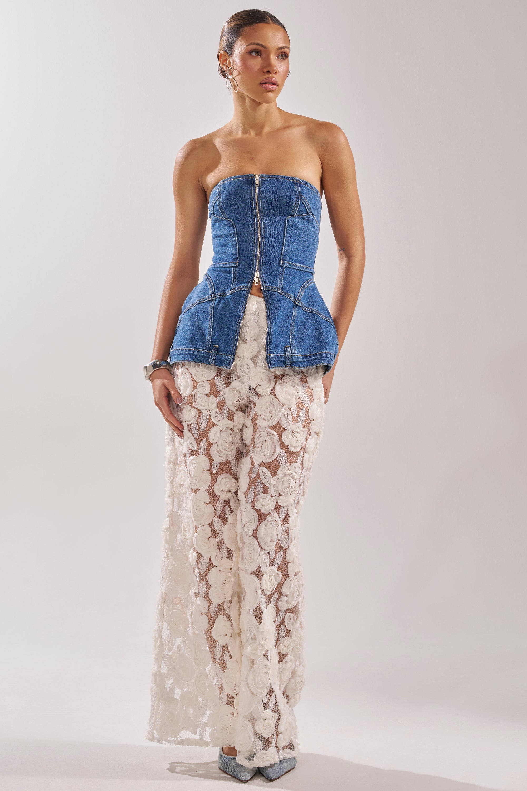 A woman models the UPSIDE DOWN DENIM CORSET, a strapless top paired with sheer white lace floral pants. Her hair is pulled back, and she accessorizes with earrings and pointed-toe shoes against a plain background. 8