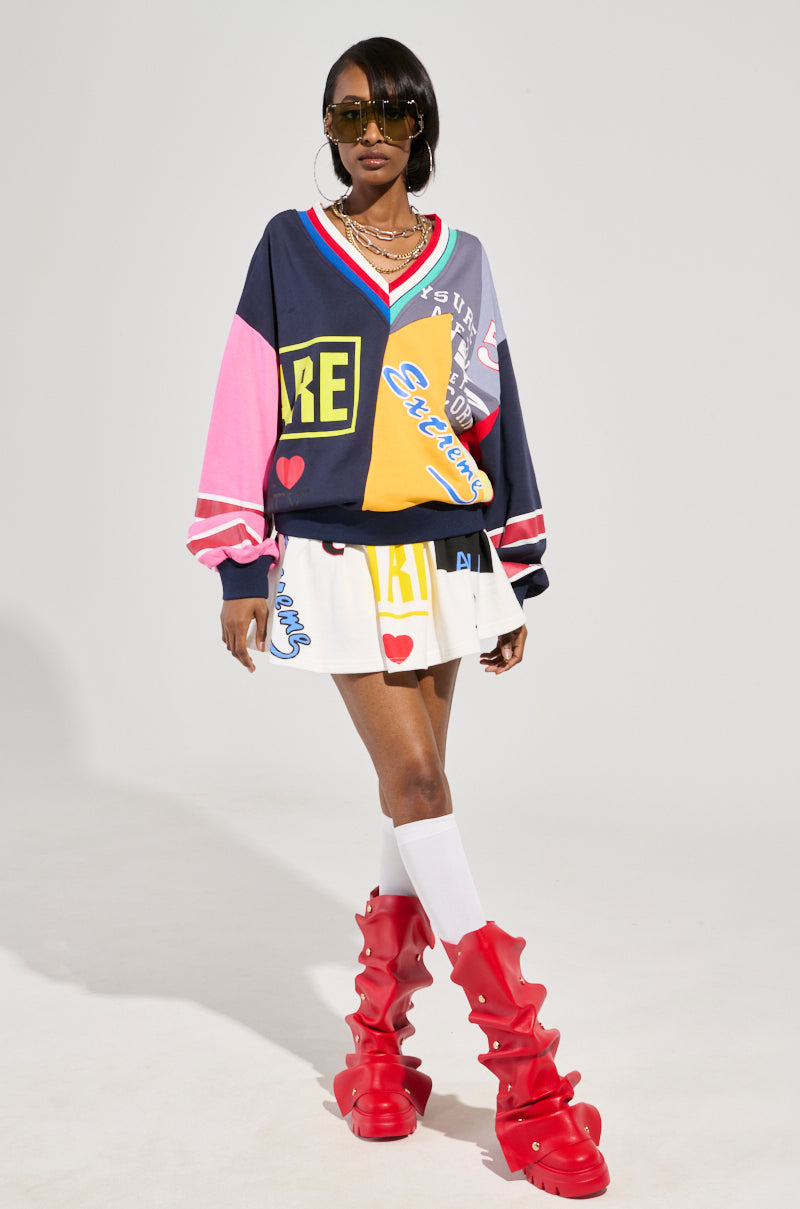 A stylish individual stands confidently in a colorful, graphic sweater and the WE ARE INFINITE PLEATED MINI SKIRT. They wear oversized sunglasses, layered necklaces, striking red wavy boots over knee-high white socks, all crafted from a cotton-blend ensemble set against a plain background.