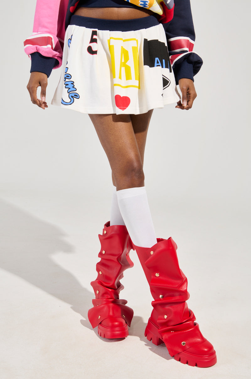 She is sporting the WE ARE INFINITE PLEATED MINI SKIRT adorned with colorful graphic designs, paired with red slouchy knee-high boots, an athletic-inspired pink and navy jacket, and long white socks. The outfit captures a bold and playful fashion sense.