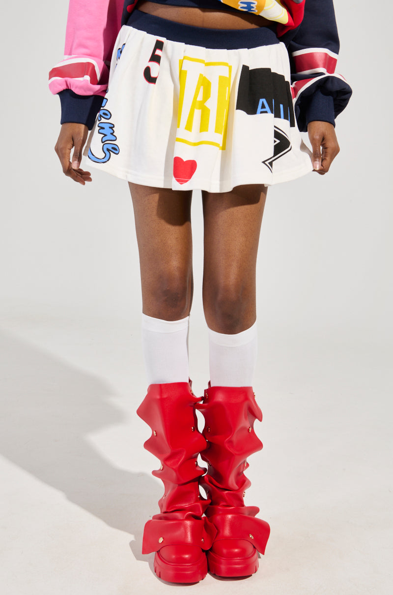 A person wearing the WE ARE INFINITE PLEATED MINI SKIRT featuring various prints, paired with a black and pink long-sleeve top made from a cotton-blend fabric, white knee-high socks, and oversized red boots with wavy designs stands against a plain background.