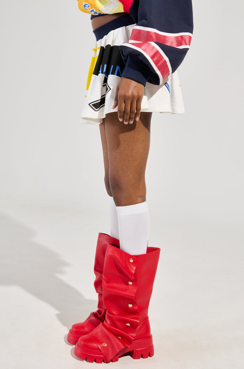 A person stands wearing the WE ARE INFINITE PLEATED MINI SKIRT, paired with an athletic-inspired dark long-sleeve shirt featuring pink and white stripes, complemented by vibrant red boots. White knee-high socks peek out from the boots, all set against a plain light background.