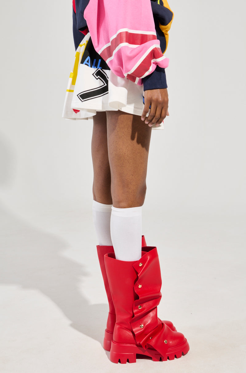 A person sporting a dynamic ensemble that includes a pink and navy top, the WE ARE INFINITE PLEATED MINI SKIRT, knee-high white socks, and vibrant red boots. The outfit showcases an avant-garde and playful style with athletic-inspired details crafted from a chic cotton-blend fabric.