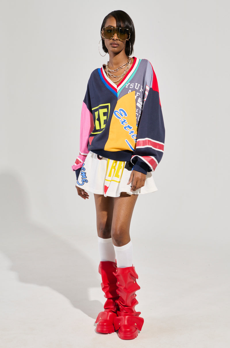 A person stands confidently wearing a colorful cotton-blend oversized sweater with graphic patterns, the WE ARE INFINITE PLEATED MINI SKIRT, and red knee-high boots. They have sunglasses and layered necklaces, posing against a plain background.