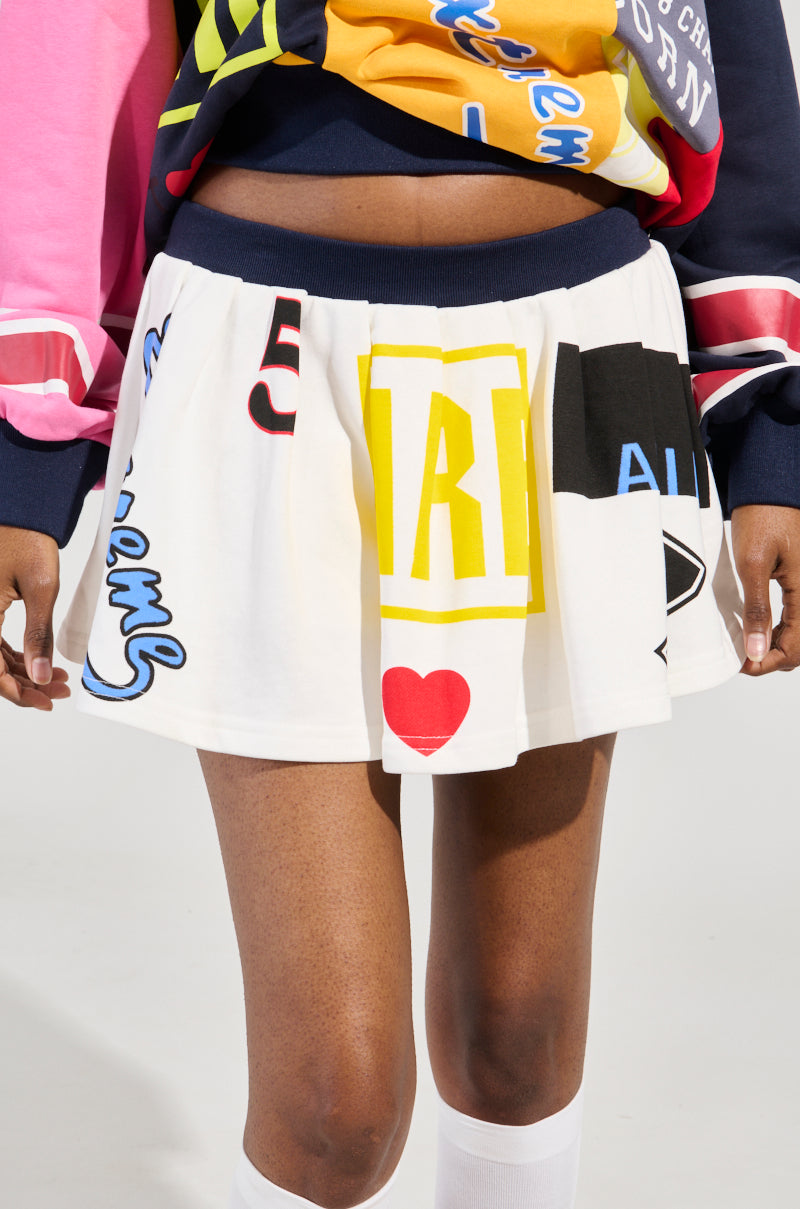 Wearing the WE ARE INFINITE PLEATED MINI SKIRT, adorned with vibrant letters, numbers, and symbols, a person stands out in their athletic-inspired ensemble. The skirt's heart and abstract designs complement a multicolored top and white socks, creating a striking contrast against the plain background.