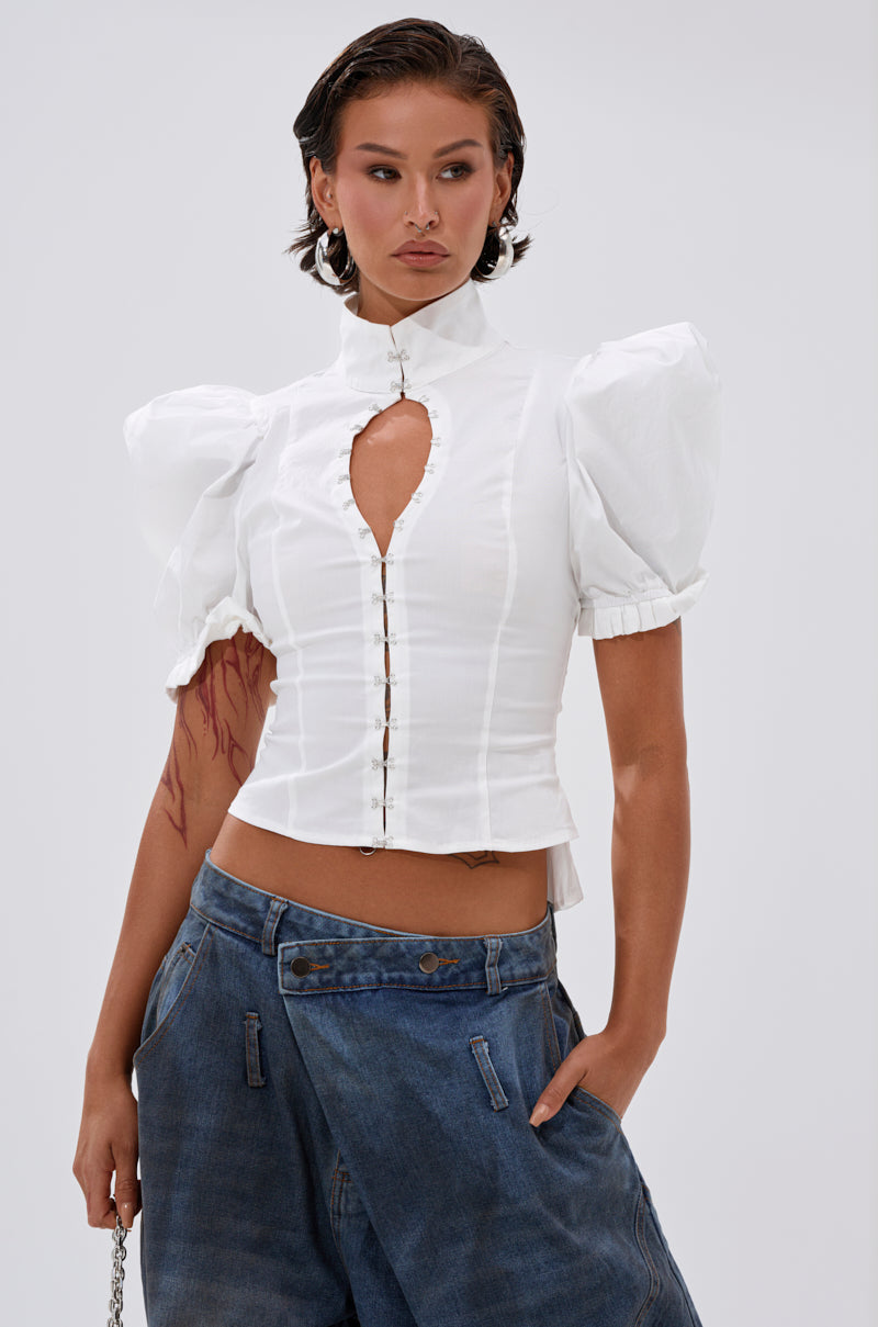 A woman poses confidently in the TOO COOL FOR YOU LACEUP BLOUSE—a fitted white puff sleeve top with a high collar and keyhole cut-out—paired with loose blue jeans. Her short, slicked-back hair adds to the modern look against a plain background.