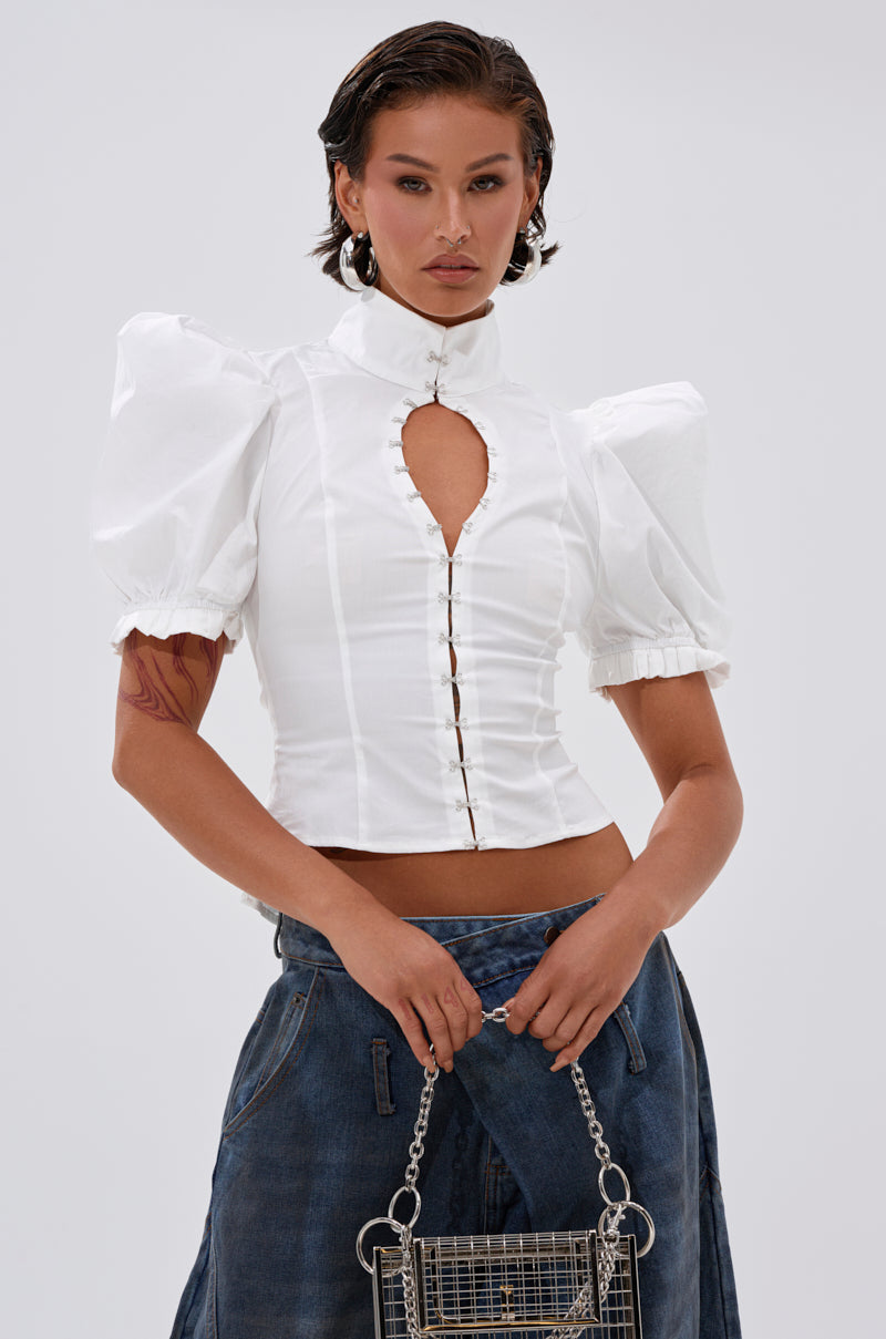 A woman wears the TOO COOL FOR YOU LACEUP BLOUSE—white, high-neck with puffed sleeves, keyhole cutout, and metallic clasps—paired with wide-leg blue jeans, a chain-strap bag, short dark hair, and silver hoop earrings.