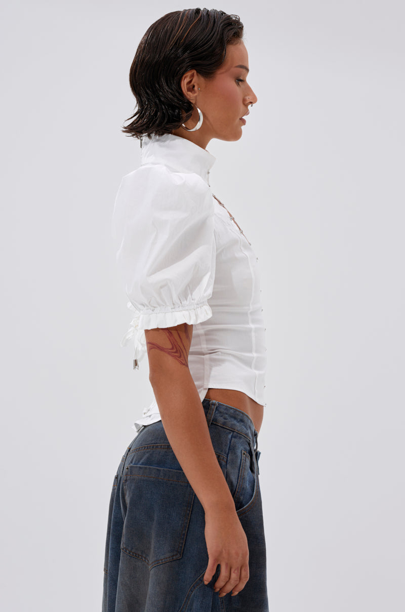 A woman with short dark hair is shown in profile, wearing the TOO COOL FOR YOU LACEUP BLOUSE with puffed sleeves and high-waisted blue jeans. She has large hoop earrings, a visible upper-arm tattoo, and stands against a plain light background.