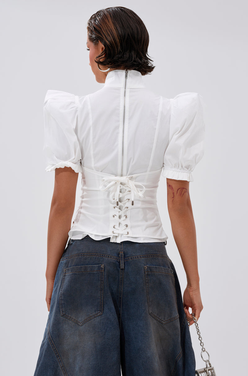 A person with short, dark wet hair is seen from behind wearing the TOO COOL FOR YOU LACEUP BLOUSE—a white corset-style top with puffed sleeves and lace-up zippered back—paired with high-waisted dark blue wide-leg jeans.