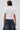 A person with short, dark wet hair is seen from behind wearing the TOO COOL FOR YOU LACEUP BLOUSE—a white corset-style top with puffed sleeves and lace-up zippered back—paired with high-waisted dark blue wide-leg jeans.