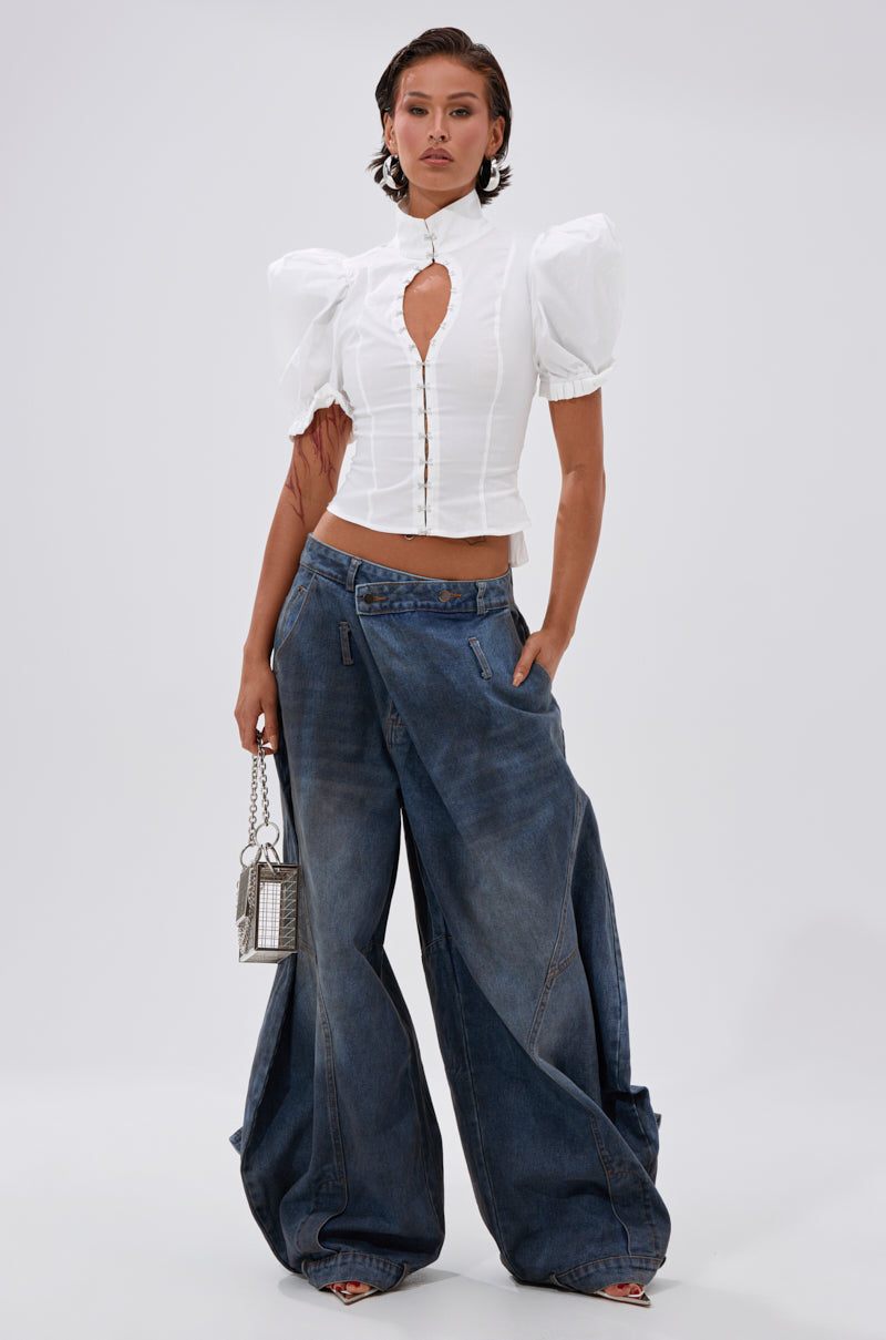 A woman poses against a plain background in the TOO COOL FOR YOU LACEUP BLOUSE, styled with wide-legged blue jeans, a small purse, slicked-back hair, and open-toe shoes.