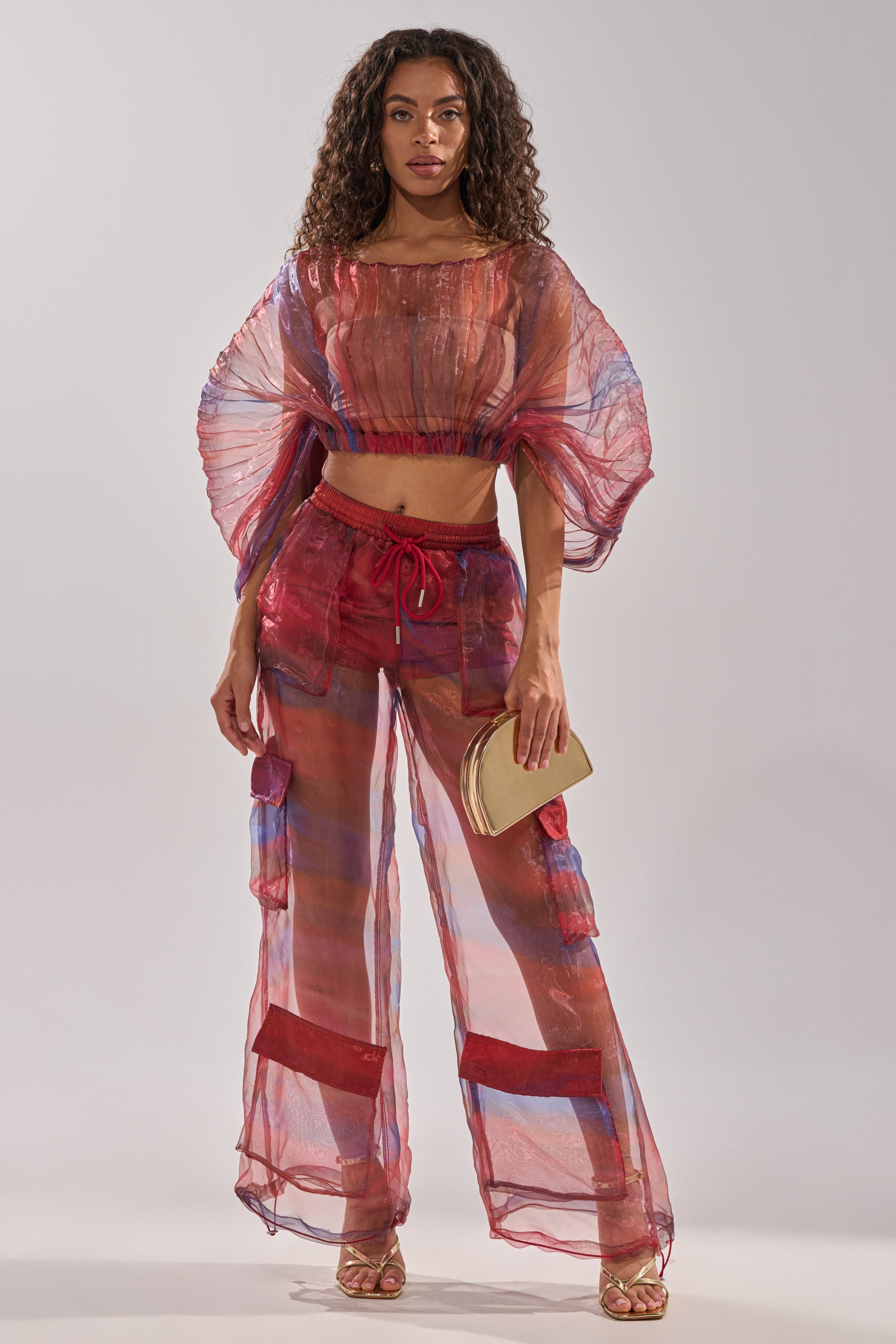 A woman stands facing forward in wide-leg LETS LIVE IN THE MOMENT CHIFFON TROUSER and a sheer colorful top over a red bikini, holding a beige clutch with gold heels and curly hair loose against a plain background. 0