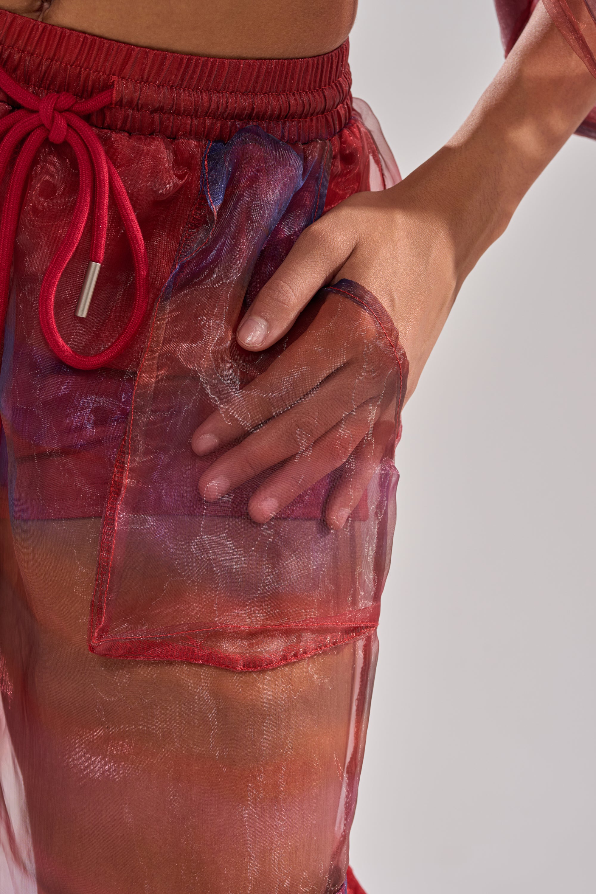 A hand rests on the sheer red pocket of the LETS LIVE IN THE MOMENT CHIFFON TROUSER, revealing skin beneath the translucent fabric. The background is plain and light-colored. 9
