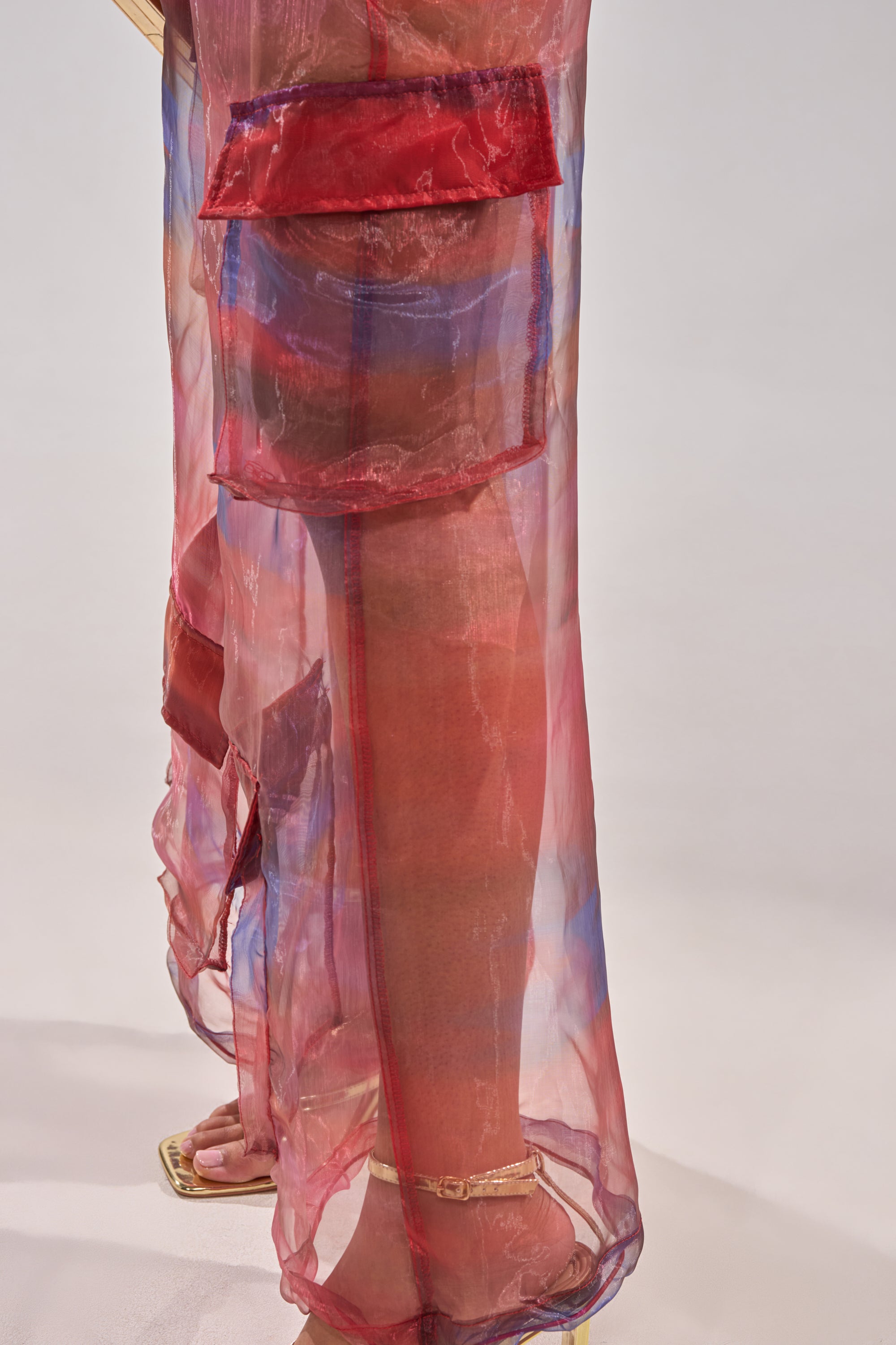 A person wears the LETS LIVE IN THE MOMENT CHIFFON TROUSER—sheer, red and blue iridescent chiffon with large cargo pockets—paired with heeled sandals, shown from the waist down against a plain light background. 10