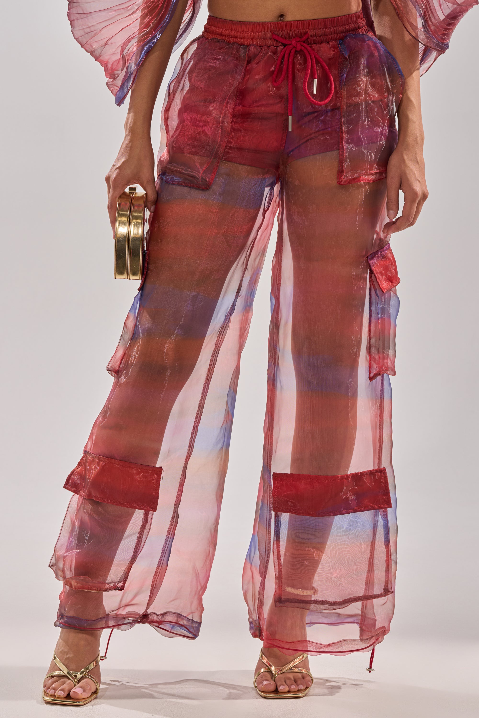 Wearing LETS LIVE IN THE MOMENT CHIFFON TROUSER—sheer, wide-leg pants with red and blue accents, large pockets, and a drawstring waist—paired with gold strappy heels and a small gold clutch. The translucent fabric reveals the legs underneath. 1