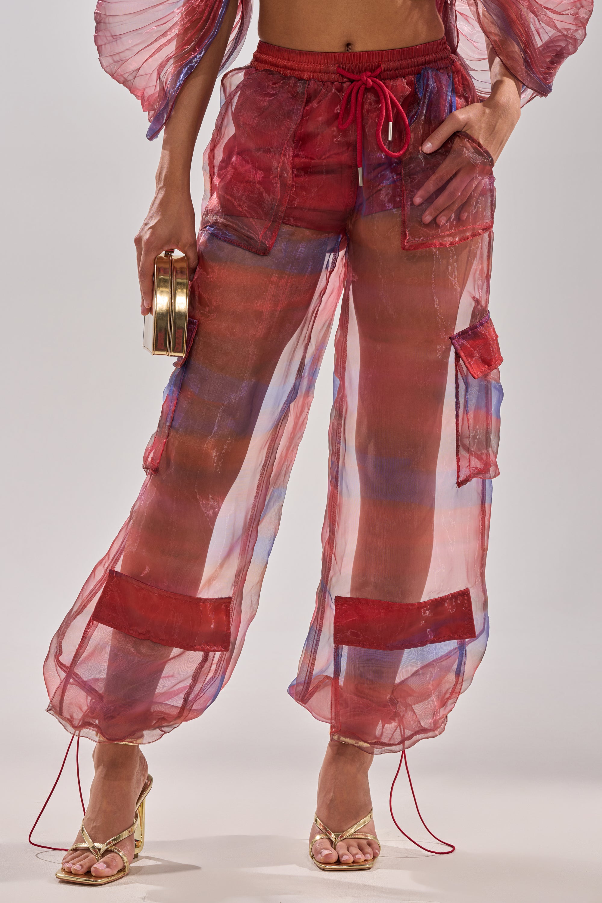 A person wears the LETS LIVE IN THE MOMENT CHIFFON TROUSER—sheer red and purple wide-leg pants with red drawstrings and square pockets—styled with a gold clutch and gold heels against a neutral background. 2