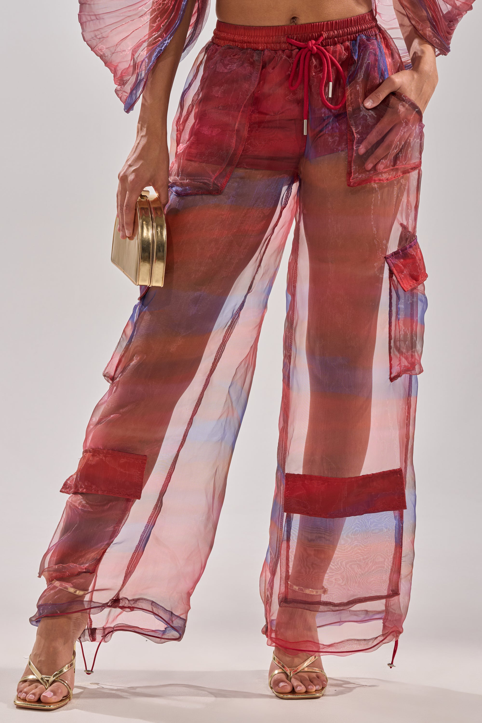 Wearing LETS LIVE IN THE MOMENT CHIFFON TROUSER—sheer, wide-leg red and blue pants with patch details, a drawstring waist, gold high-heeled sandals, and a gold clutch. 3