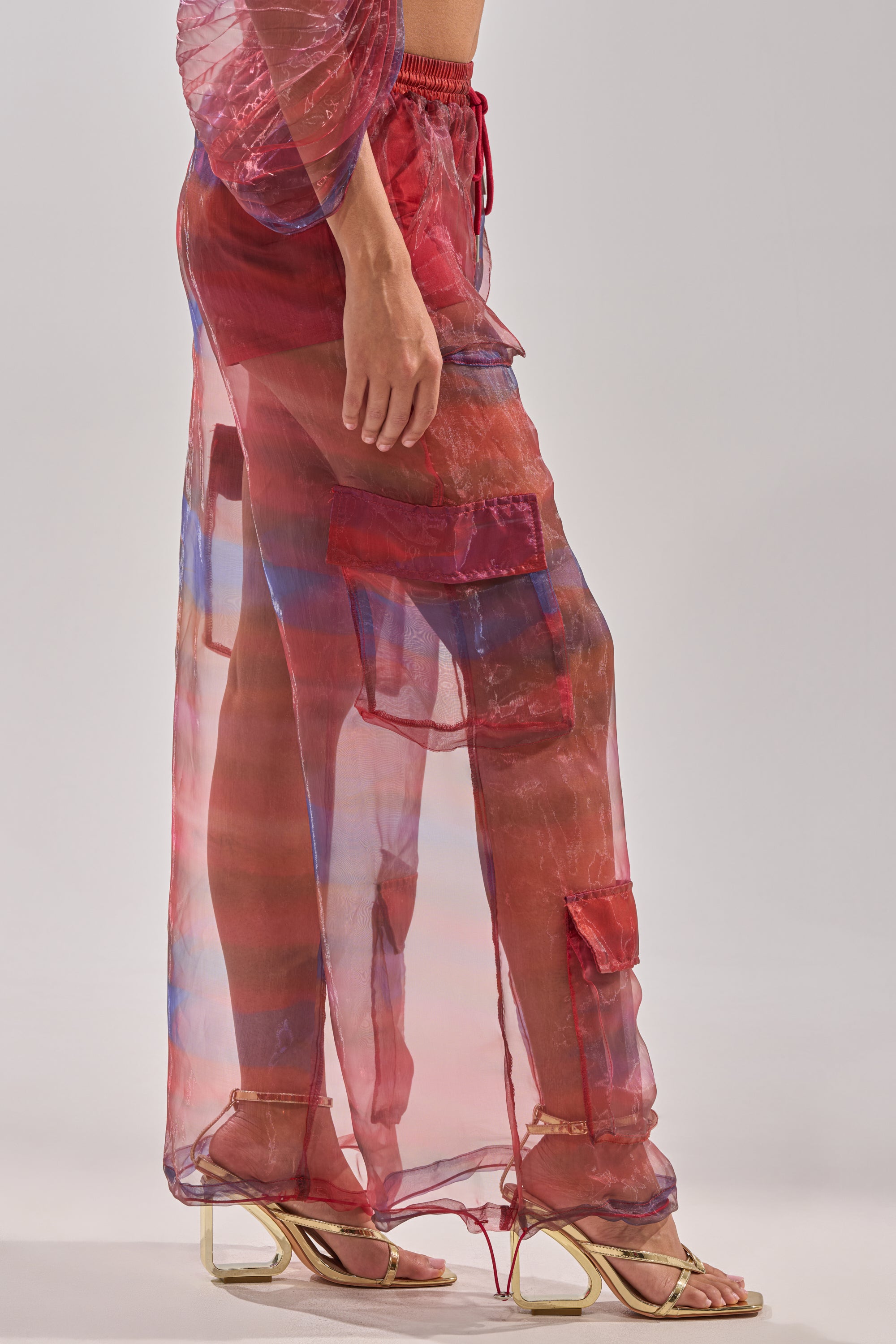 A person stands in profile wearing the LETS LIVE IN THE MOMENT CHIFFON TROUSER, sheer red wide-leg pants with patchwork details and visible red shorts underneath, styled with gold strappy heels against a plain light backdrop. 4