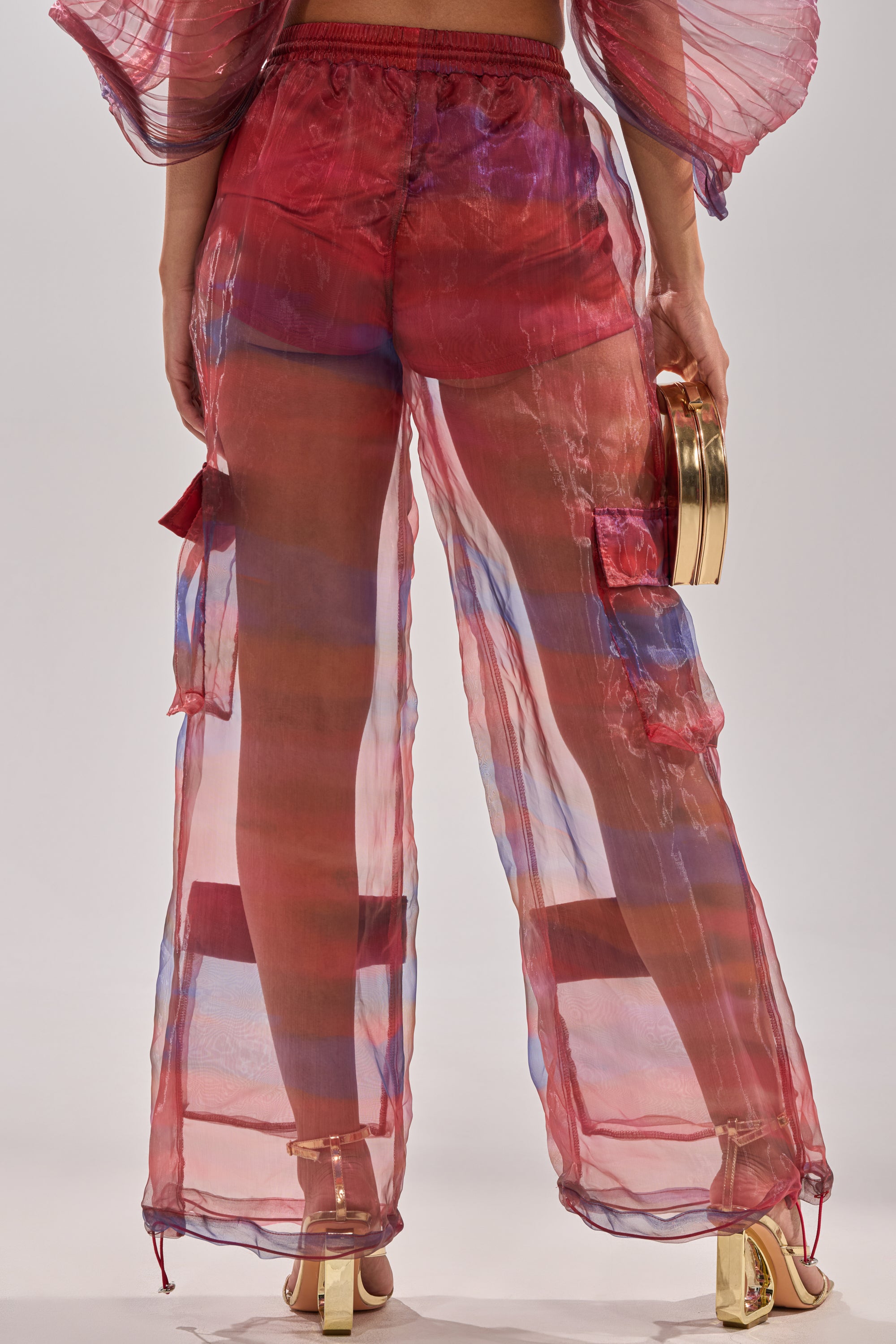 A person wears LETS LIVE IN THE MOMENT CHIFFON TROUSER in sheer, colorful chiffon over red shorts, paired with gold heels and a small gold handbag. The photo is taken from behind, highlighting the outfit’s details. 5