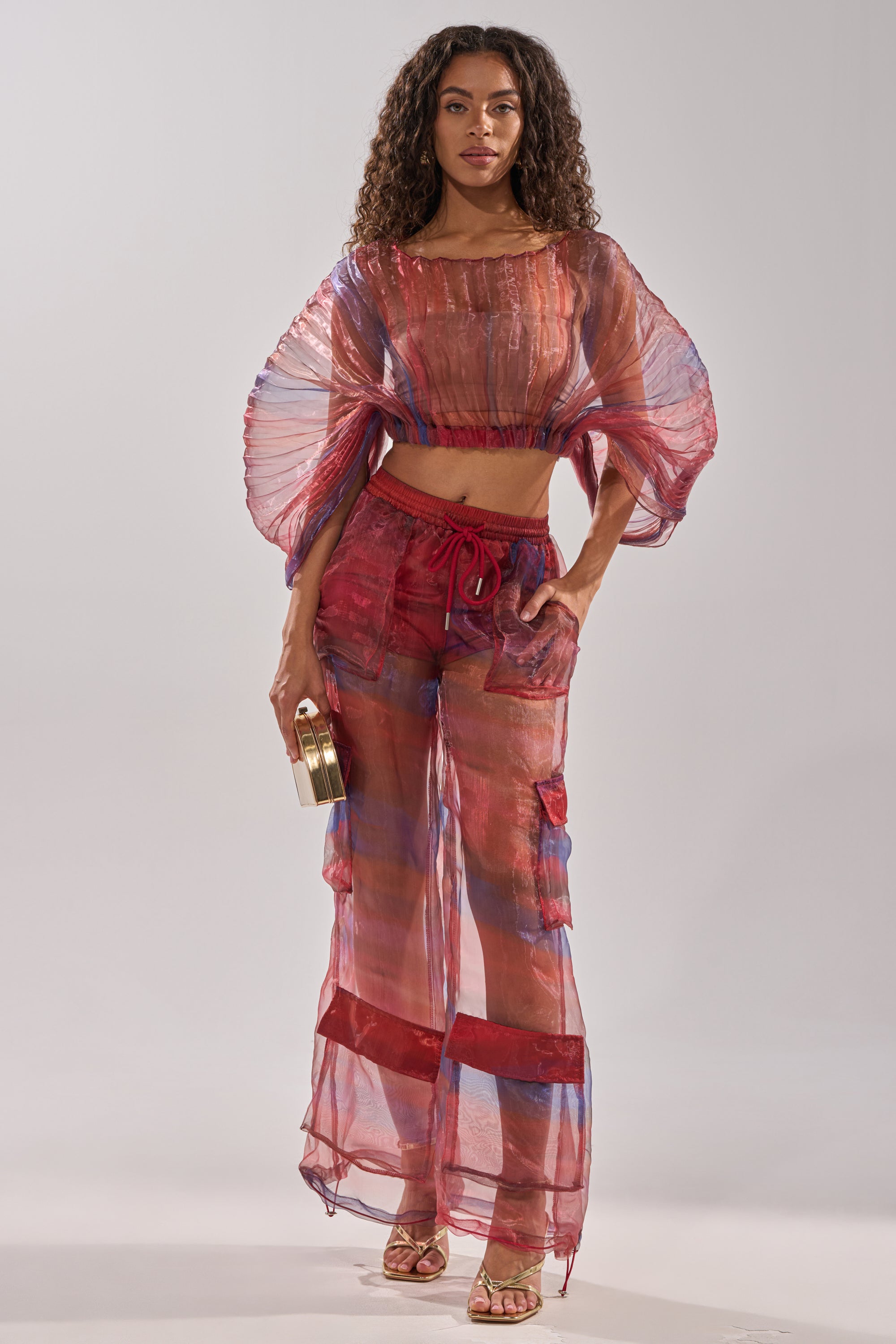 A woman wears the LETS LIVE IN THE MOMENT CHIFFON TROUSER, a sheer red and blue two-piece with wide sleeves and chiffon wide-leg pants, paired with strappy heels and a gold clutch, standing against a plain light background. 6