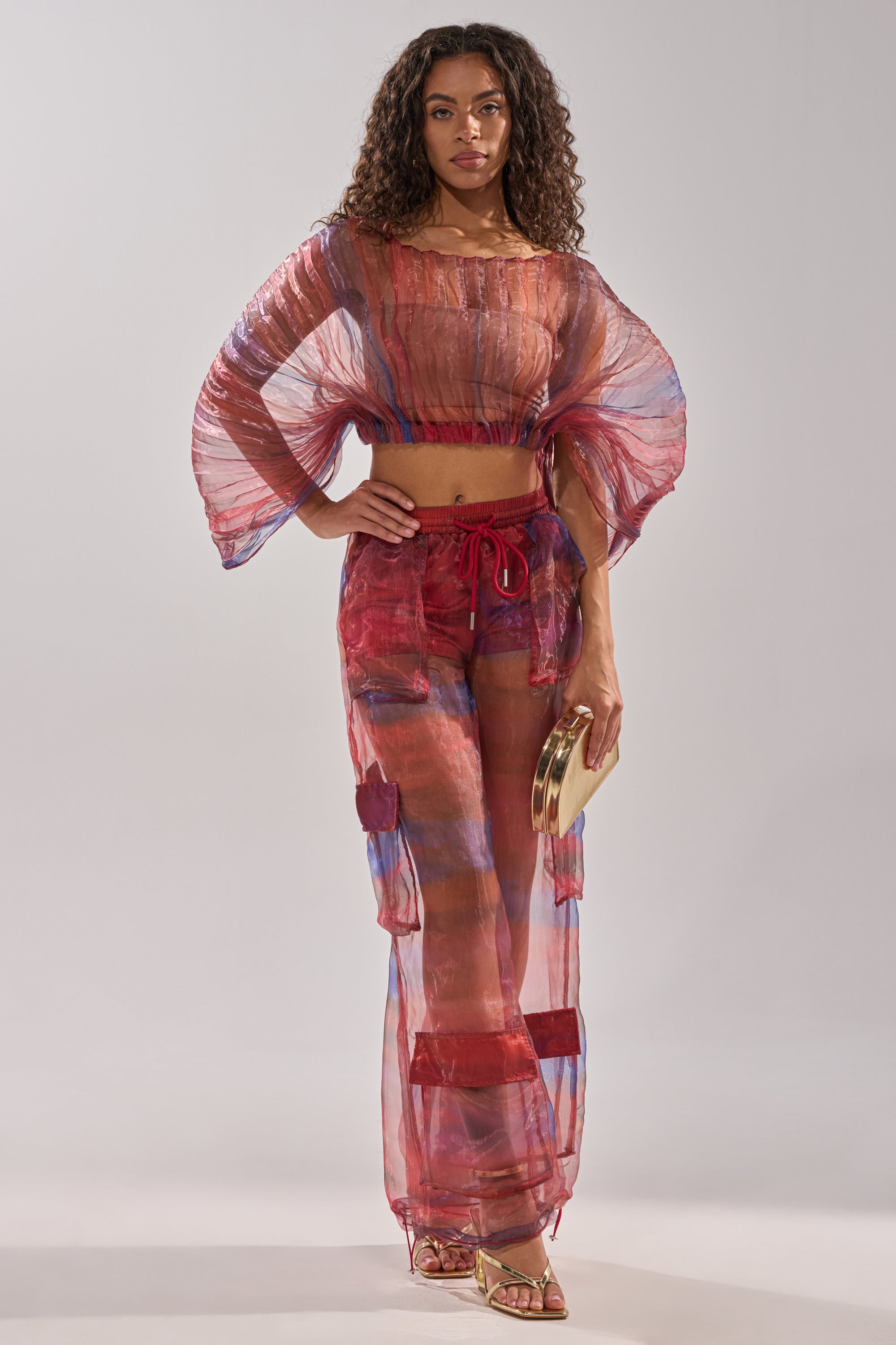 A woman wears the LETS LIVE IN THE MOMENT CHIFFON TROUSER, part of a sheer red and blue two-piece with wide sleeves. She pairs it with a small round clutch, gold sandals, and lets her loose curls frame her face. 7