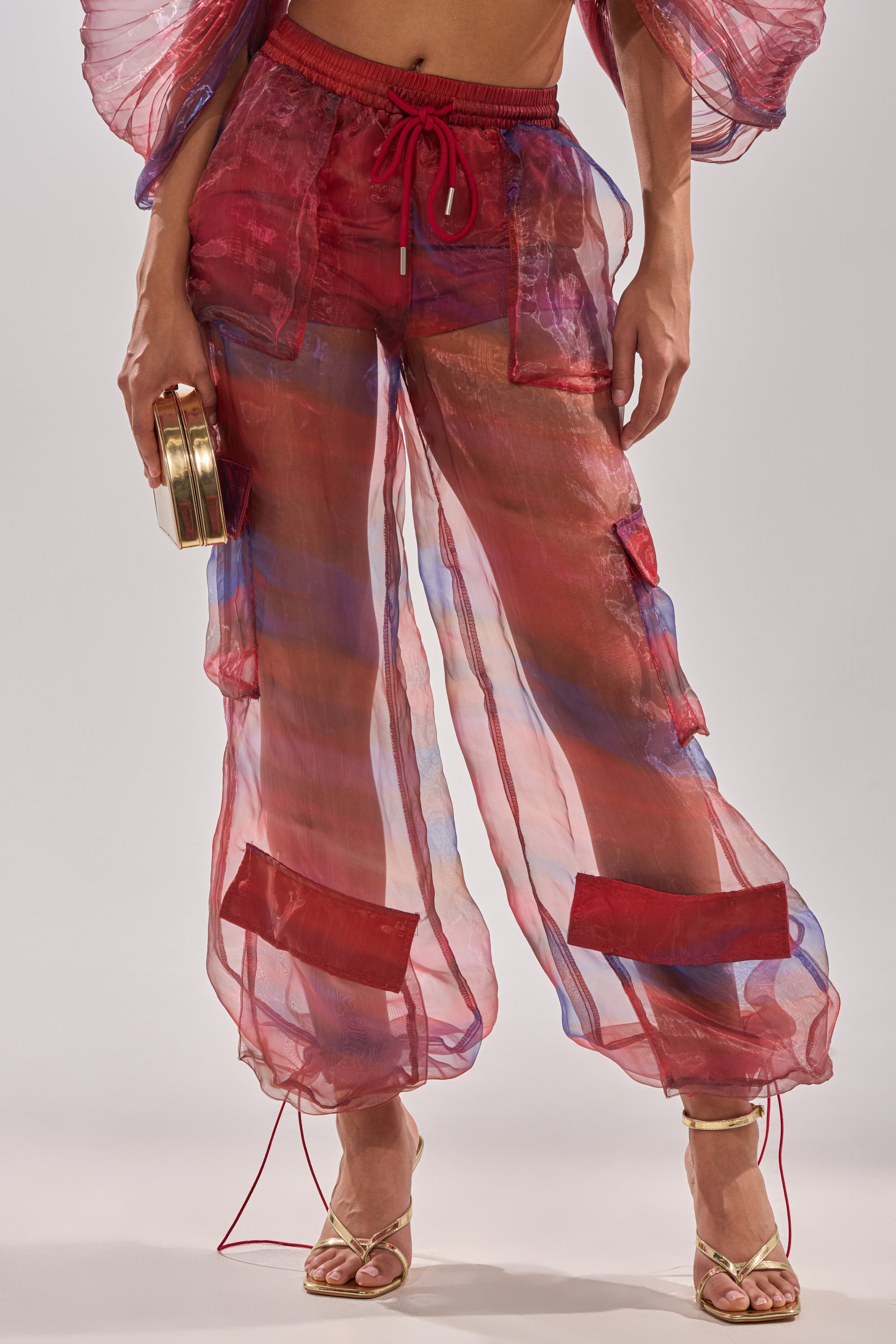 A person wears the LETS LIVE IN THE MOMENT CHIFFON TROUSER—sheer red wide-leg trousers with blue and purple hues, a red drawstring waist, and rectangular ankle patches—paired with gold strappy heels and a gold clutch. 8