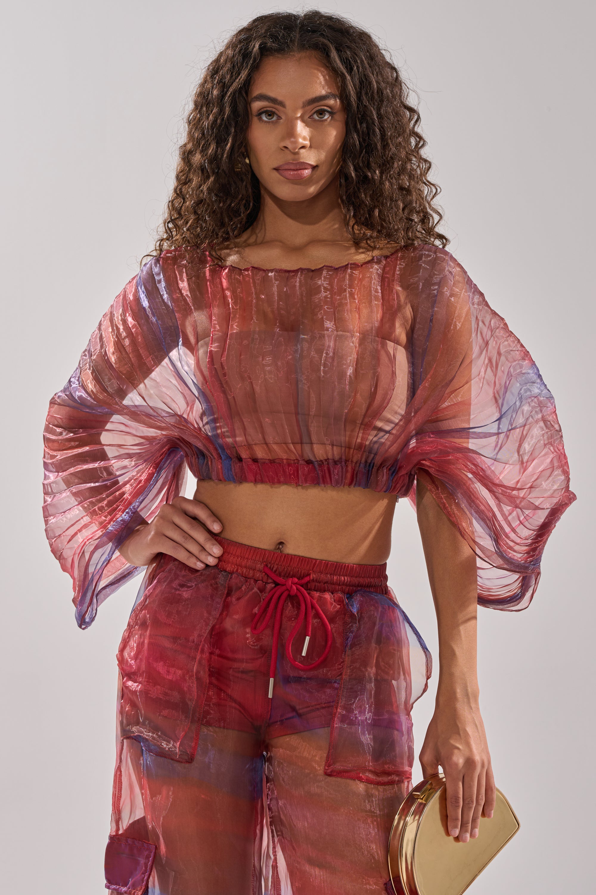 A woman with long curly hair models the LETS LIVE IN THE MOMENT CHIFFON BLOUSE—a sheer, multicolor organza top paired with matching pants over a red bralette and shorts. She holds a gold clutch and faces the camera against a plain background. 0
