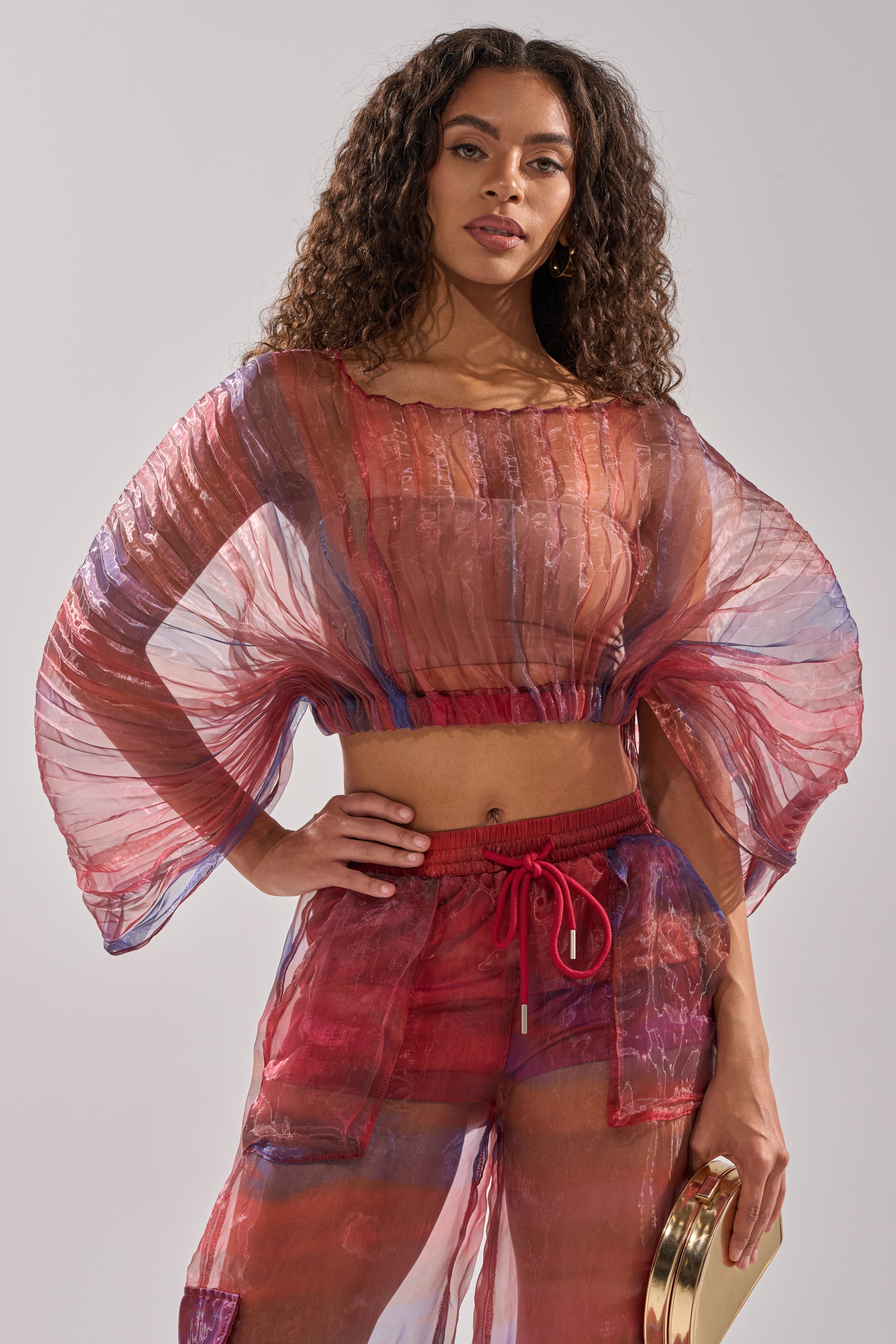 A woman with long curly hair models the LETS LIVE IN THE MOMENT CHIFFON BLOUSE—a sheer, flowy, multicolor top with wide sleeves—paired with matching pants and a gold clutch, posing confidently against a plain background. 1