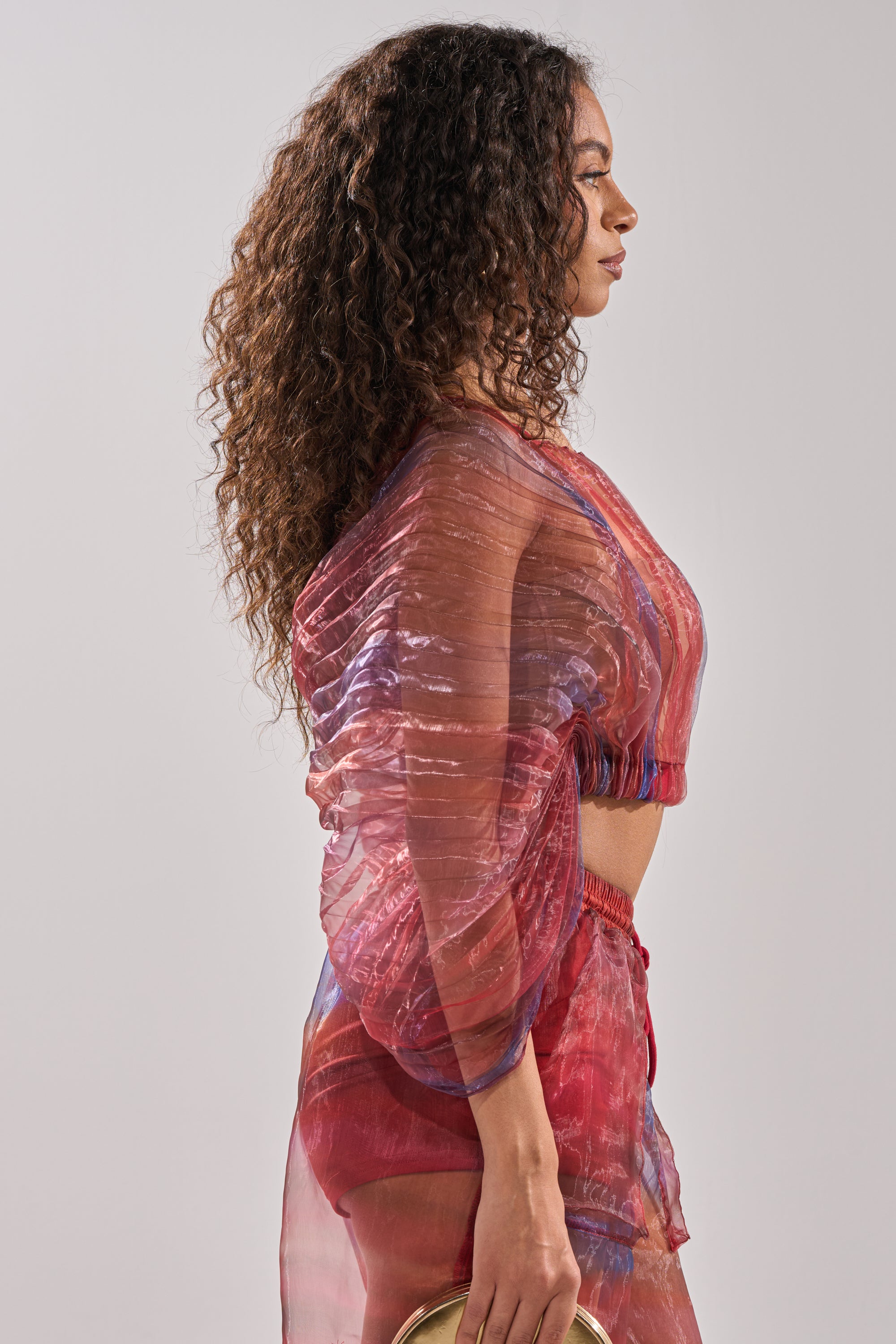 A woman with long curly hair stands in profile, wearing the LETS LIVE IN THE MOMENT CHIFFON BLOUSE in pink and purple tones. She holds a small round object in her left hand against a plain light background. 2